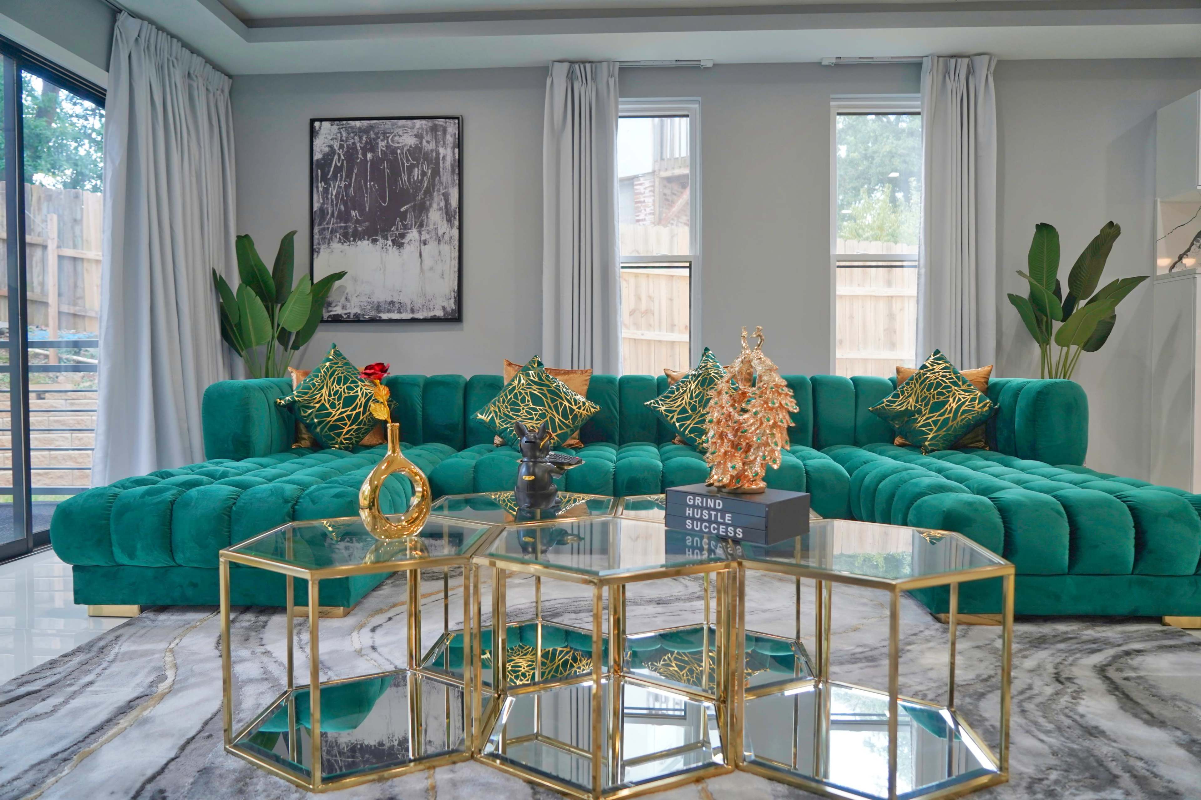 A modern living room features a large teal sectional sofa adorned with patterned cushions, a glass and gold hexagonal coffee table, and decorative plants, set against light-colored walls.