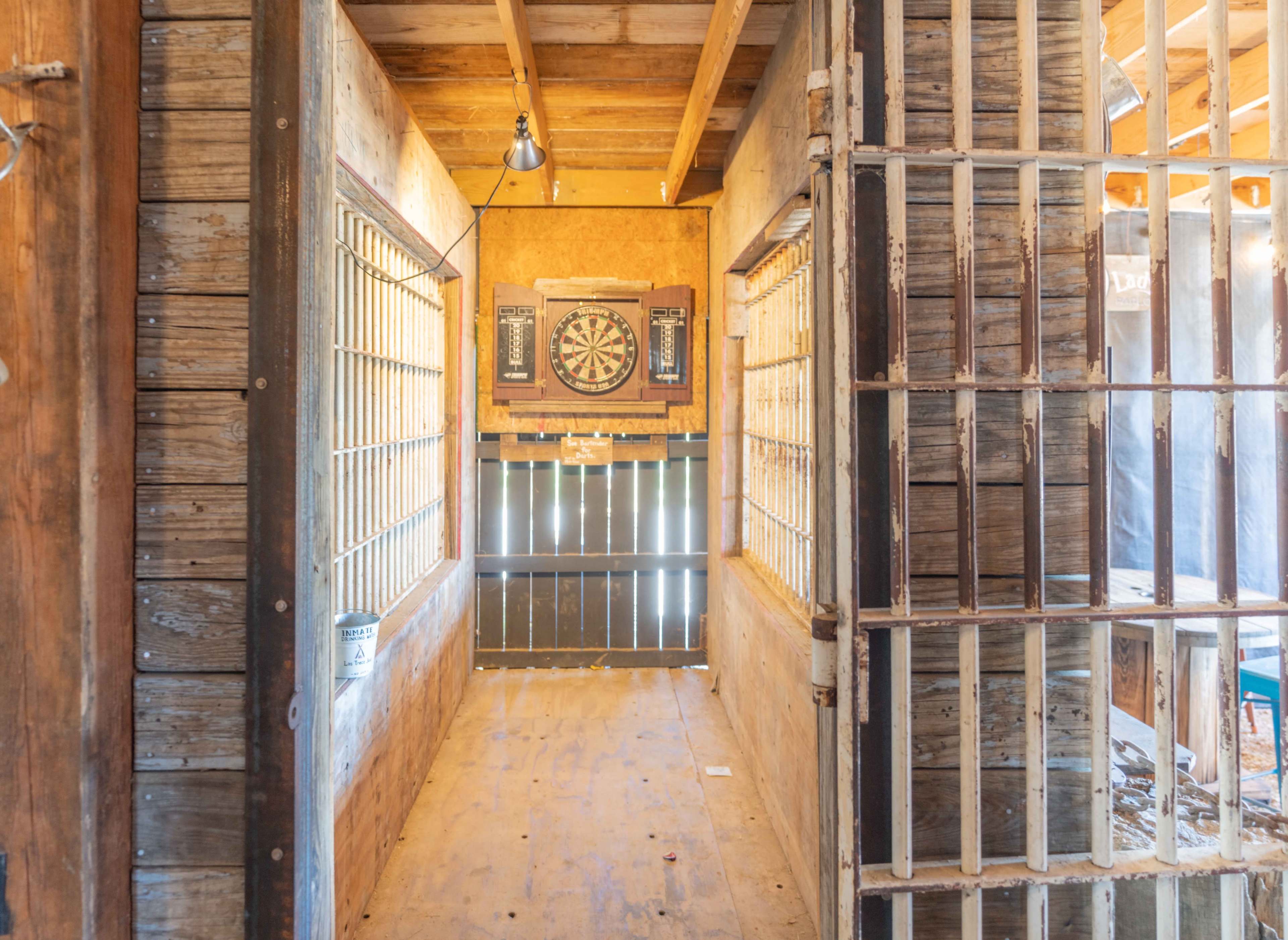 The image shows a narrow hallway between two barred cells, with a dartboard mounted on the wall at the far end.