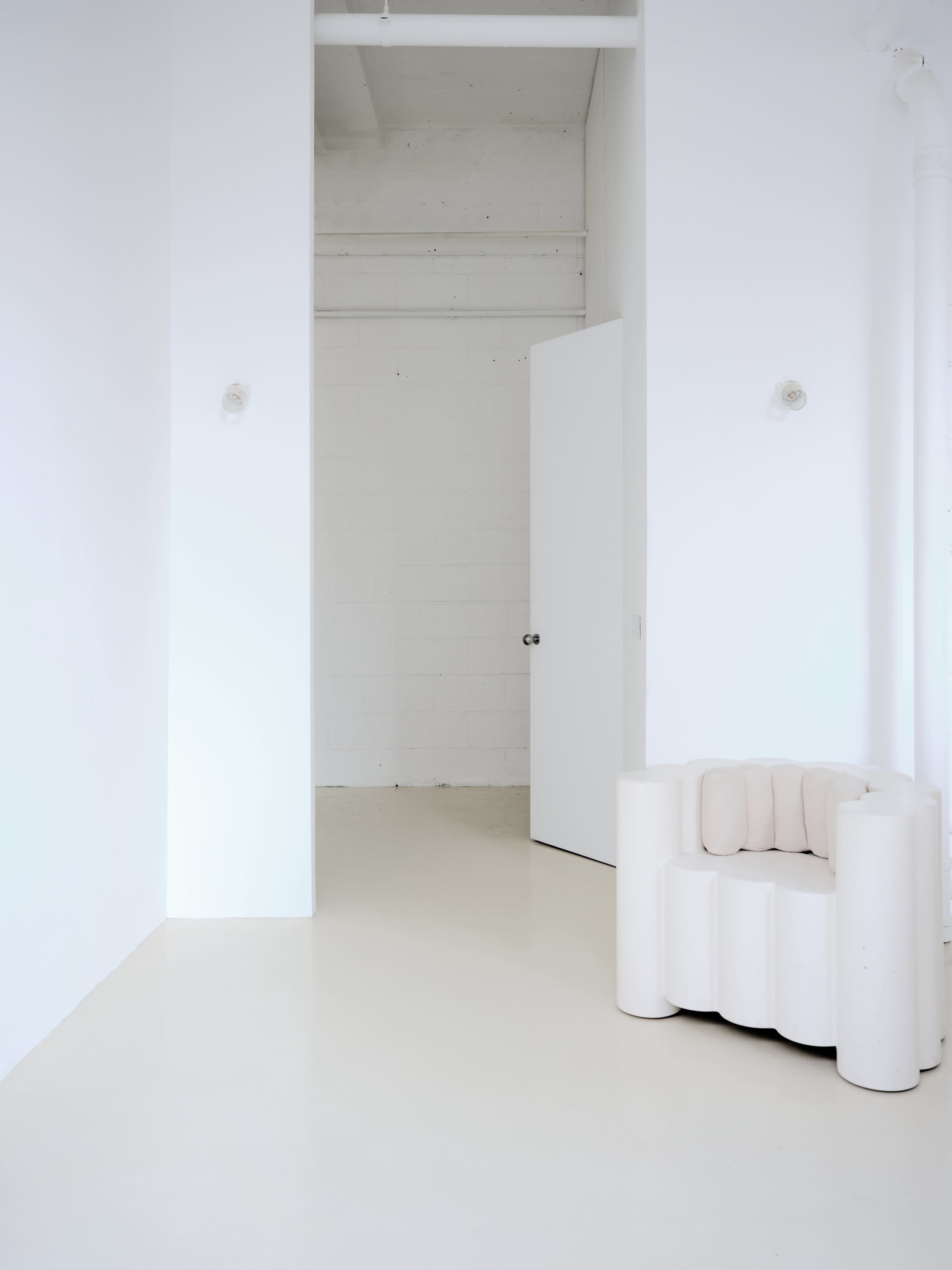A minimalist interior features a white room with a curved, white chair and an open door leading to an empty space.