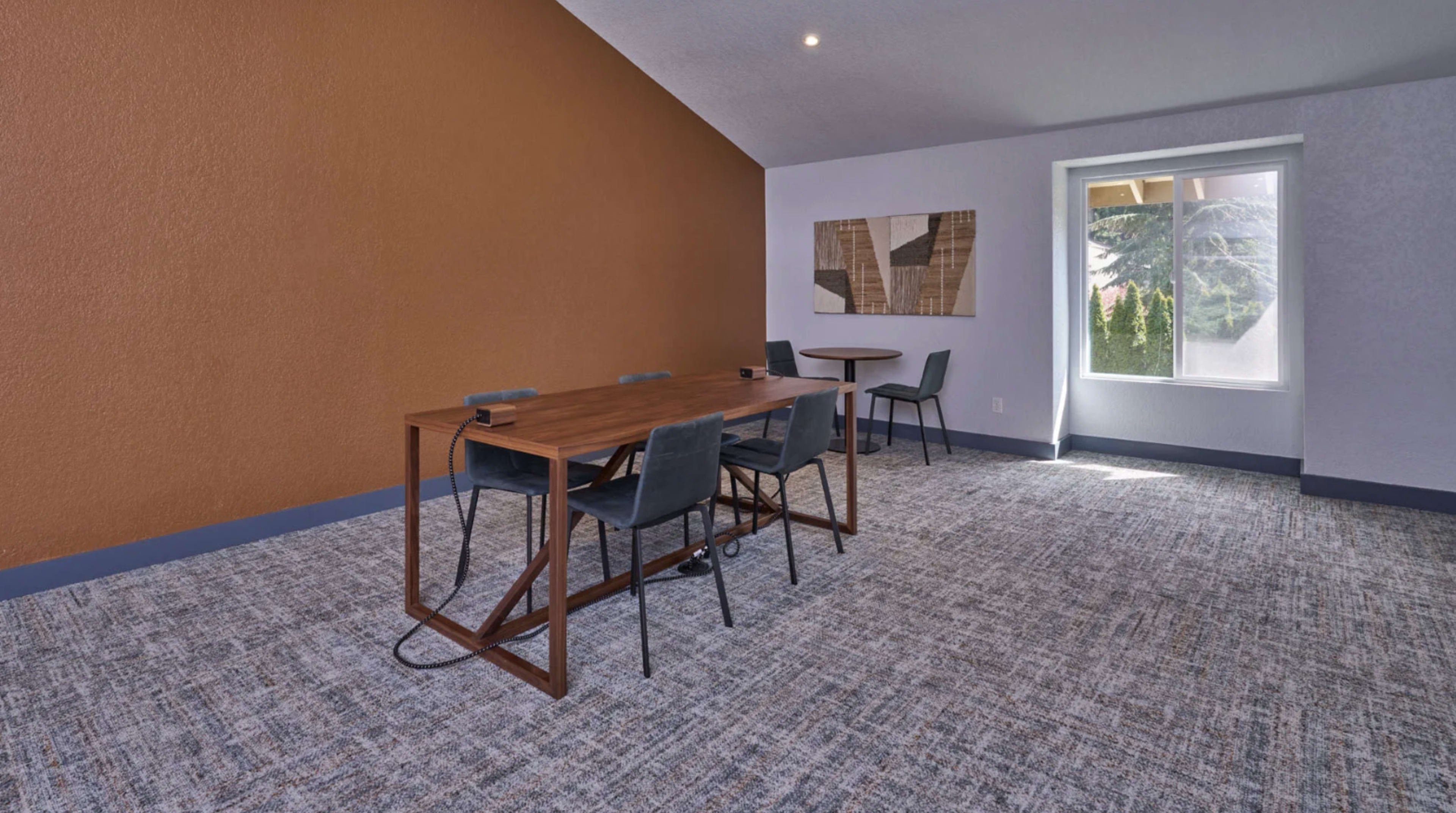 The image shows a spacious room with a wooden table surrounded by chairs, a small circular table in the background, and a large window letting in natural light.