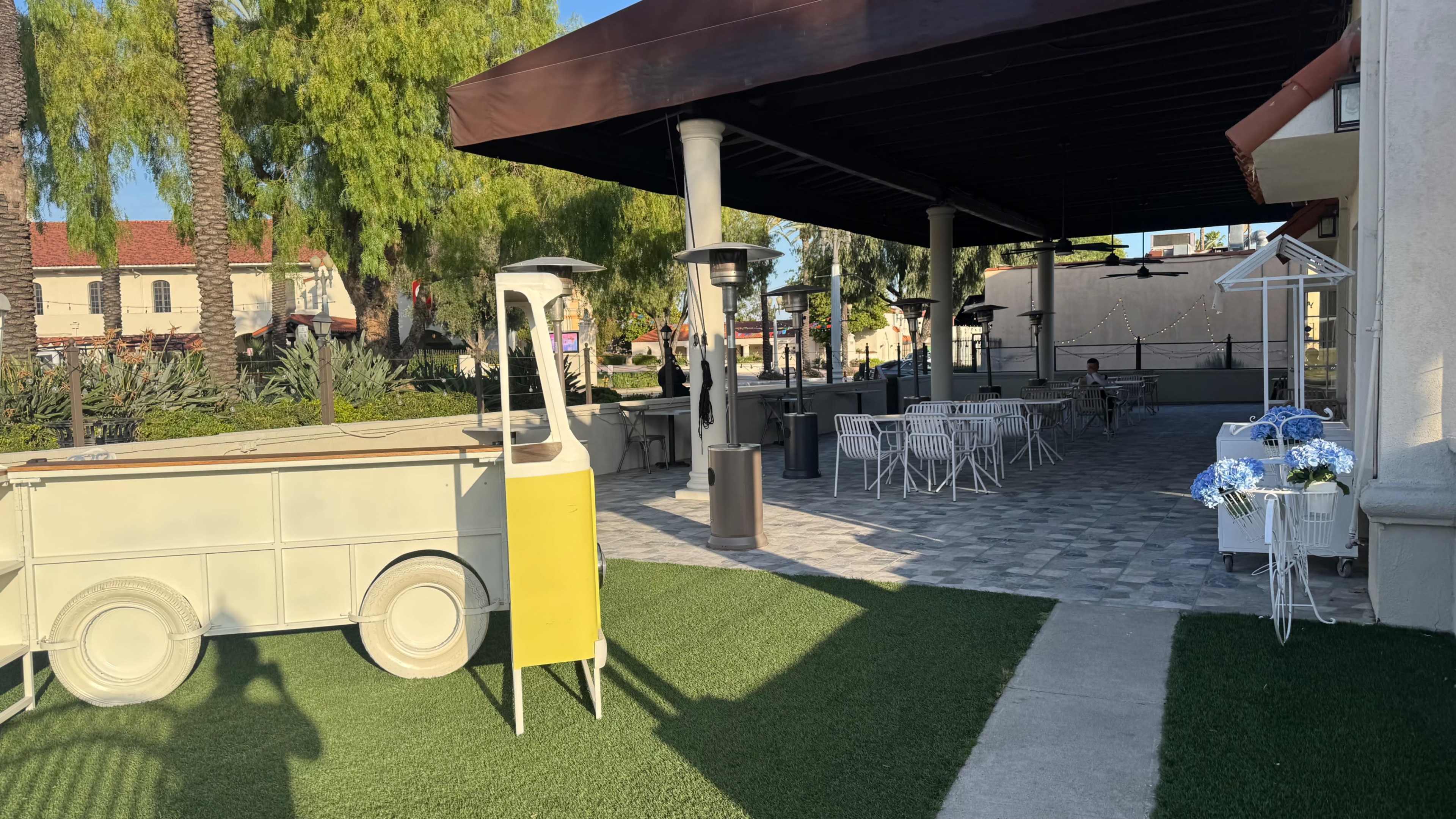 The image shows an outdoor patio area of a restaurant with a yellow cart, seating arrangements, and heating lamps under a shaded structure.