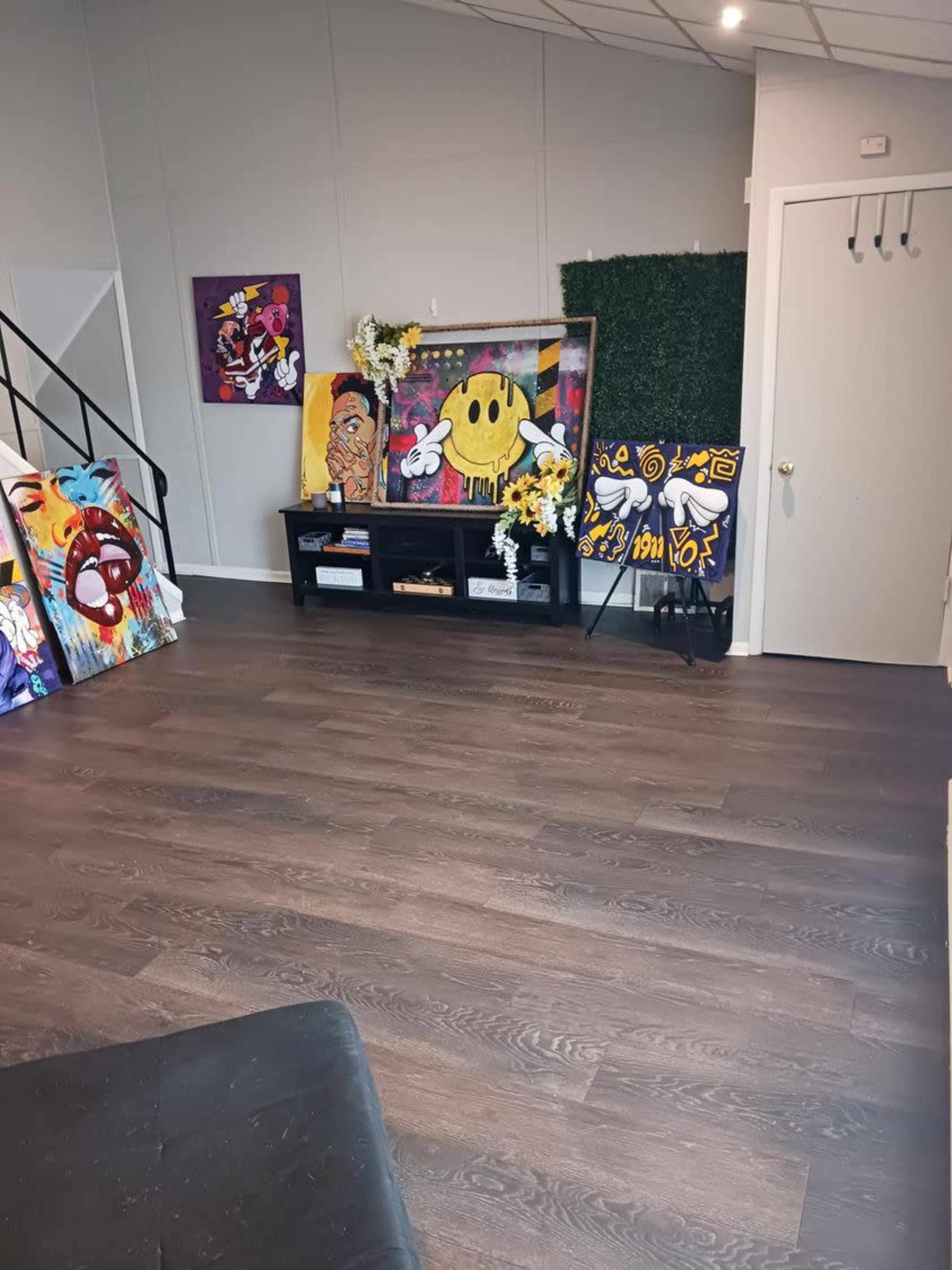 An interior room featuring a wooden floor, a display of colorful artwork on an easel, and a black console table decorated with flowers and plants.