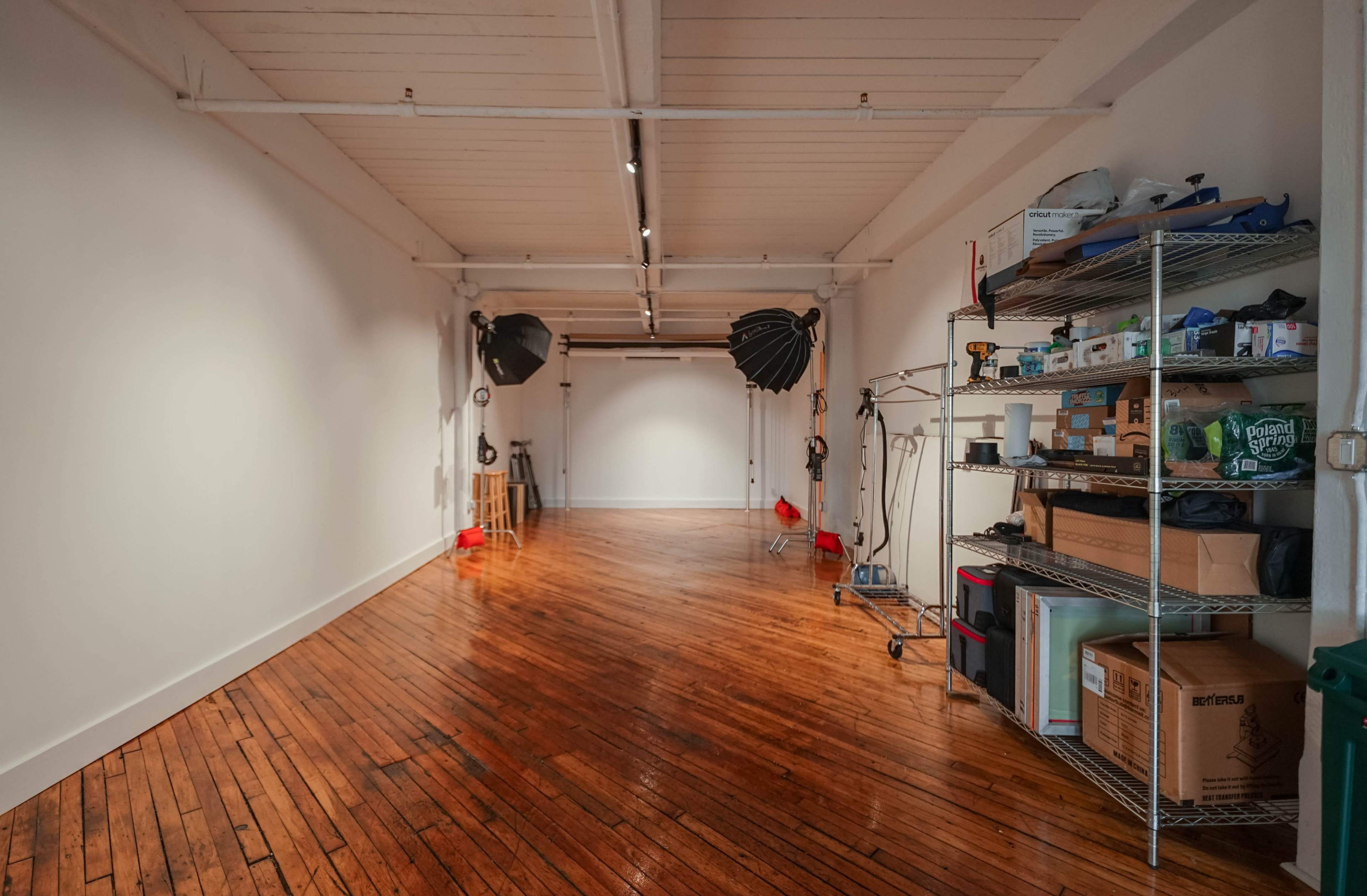 A spacious, well-lit room with polished wooden floors, two large softbox light setups, and various photography equipment stored on shelves.