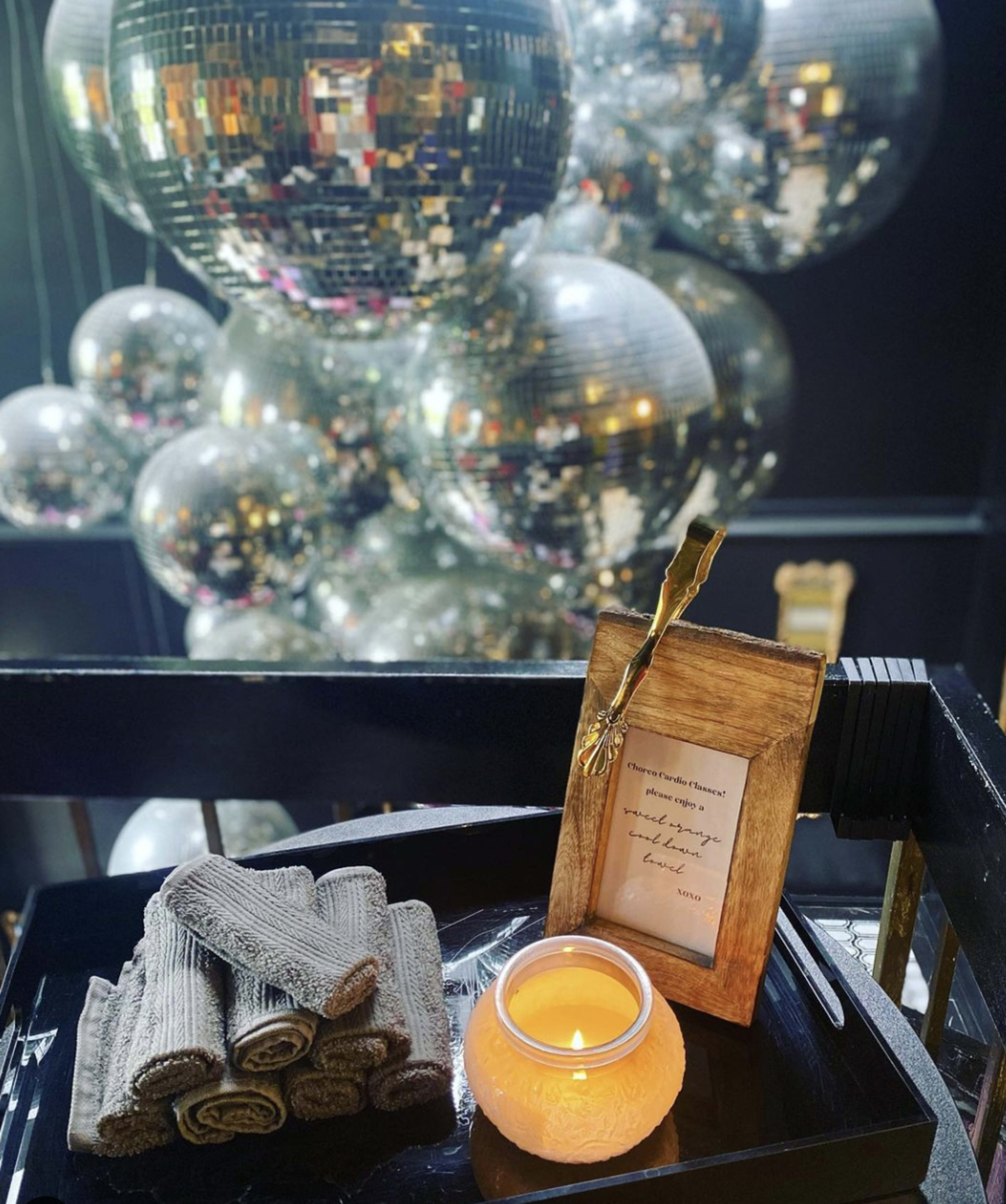 A wooden tray holds neatly rolled towels, a lit candle, and a framed note, with large disco balls reflecting light in the background.