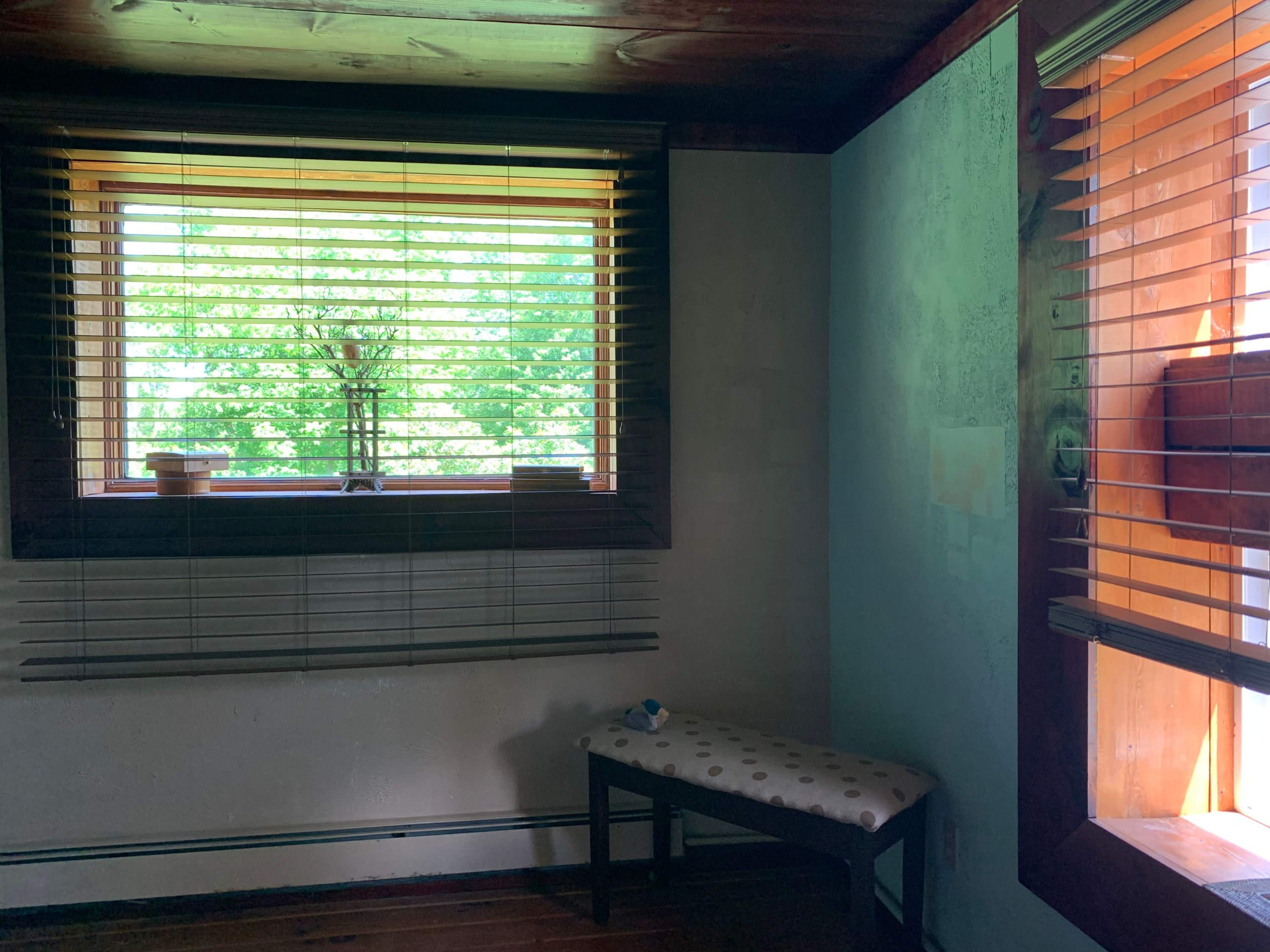 A room with two windows, one featuring blinds and a bench below, and the other partially open, allowing light to enter.