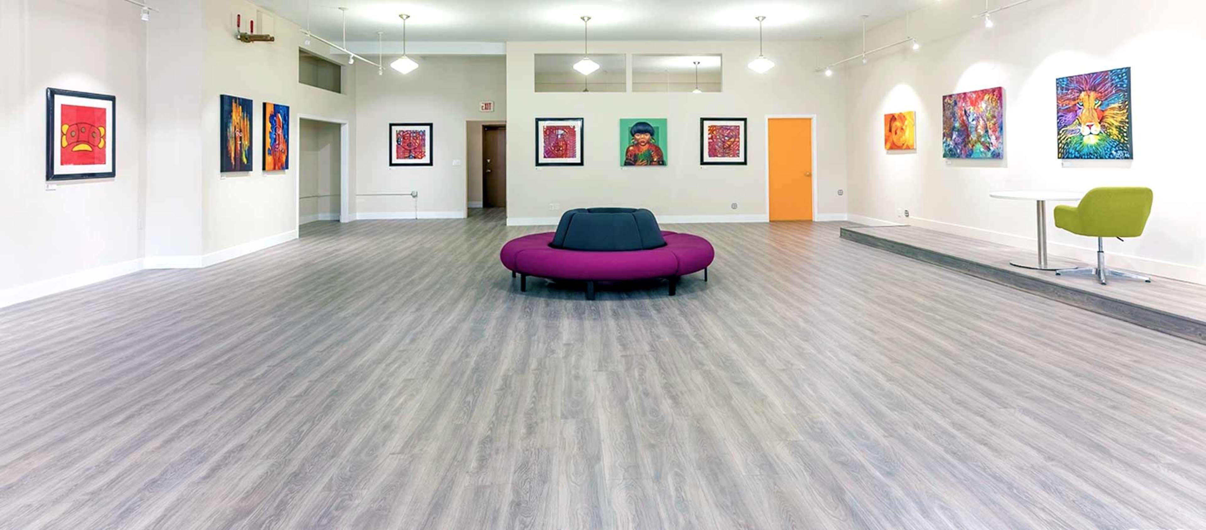 A brightly lit gallery space with a large purple seating area in the center and colorful artworks displayed on the walls.