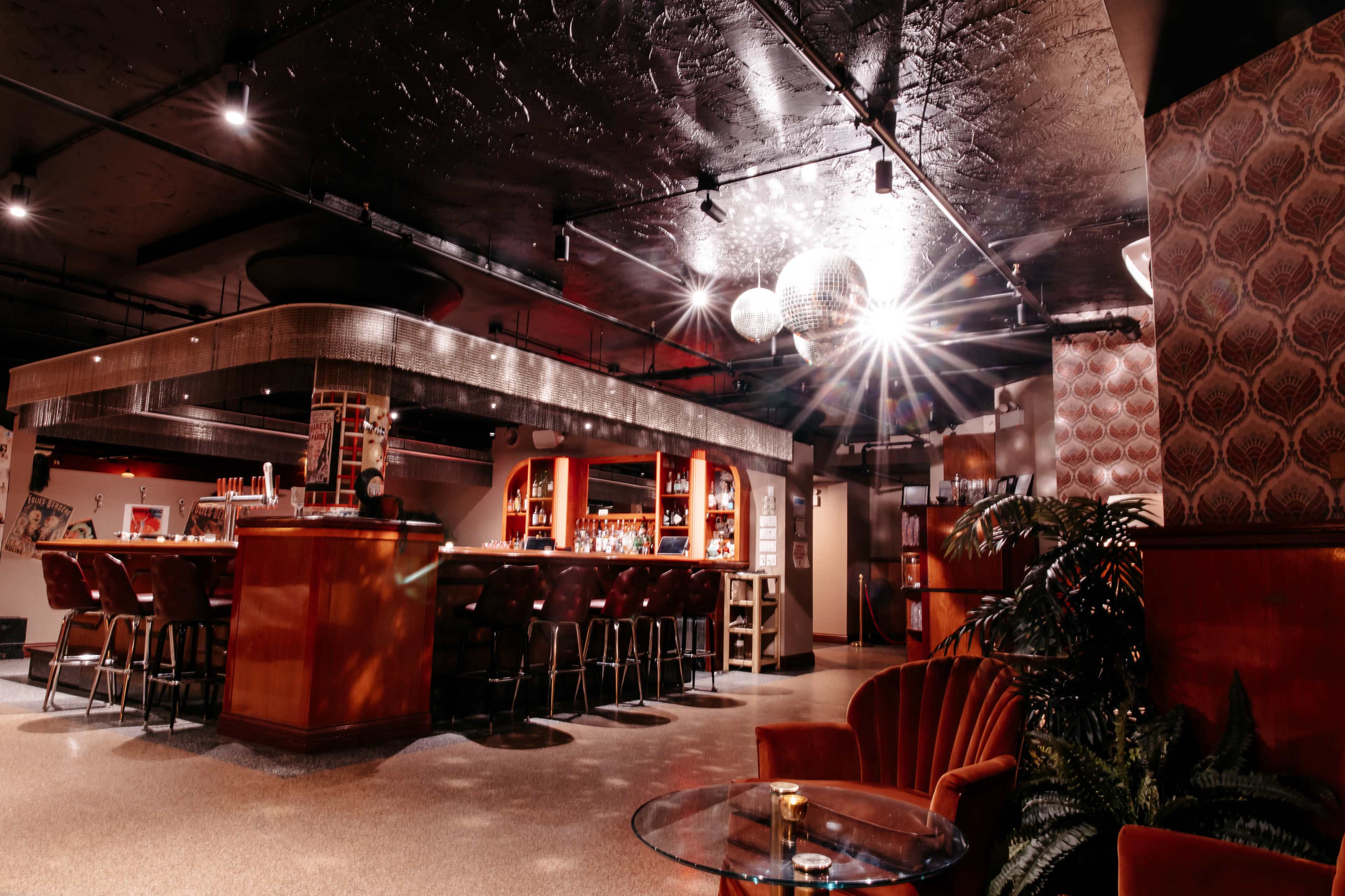 A dimly lit bar features a circular counter with high stools, surrounded by retro decor and a large disco ball reflecting light.