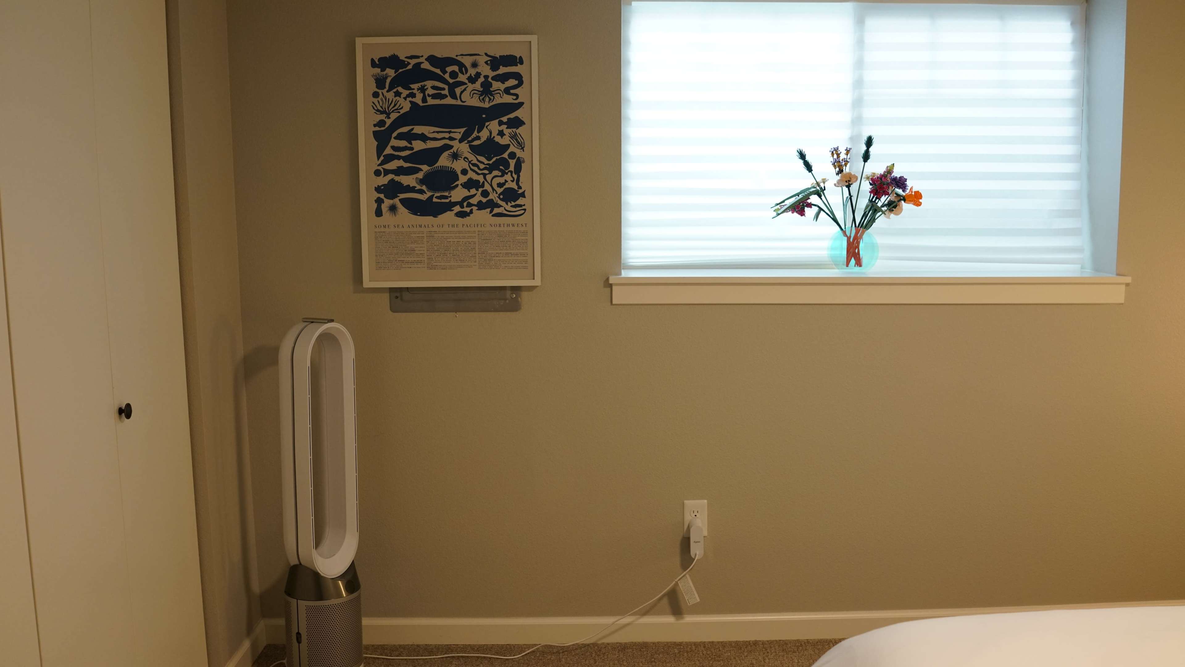 A room features a window with sheer blinds, a wall-mounted artwork, a vase of flowers on the windowsill, and a standing fan beside the bed.