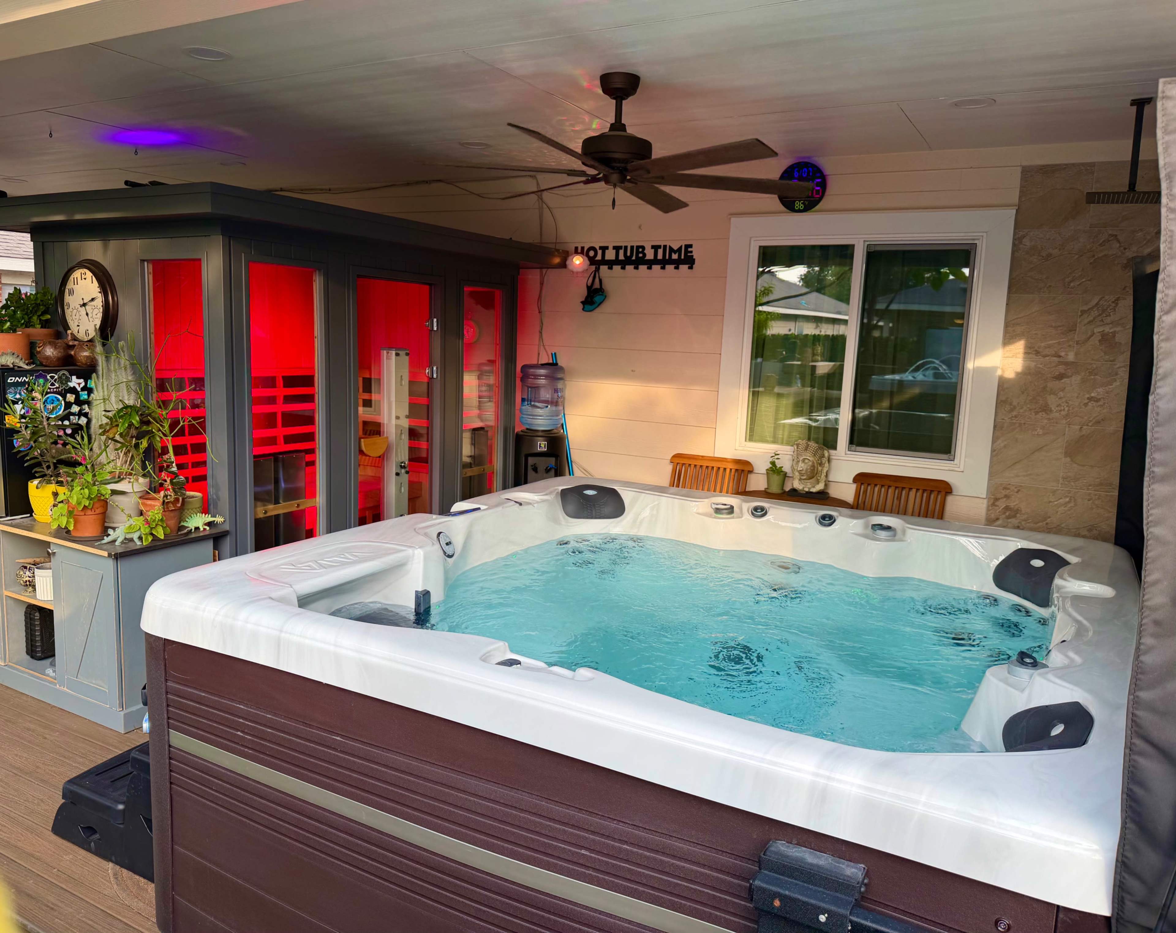 The image shows a hot tub surrounded by a wooden deck, with a nearby outdoor seating area and a cabinet featuring colorful decor and a clock.