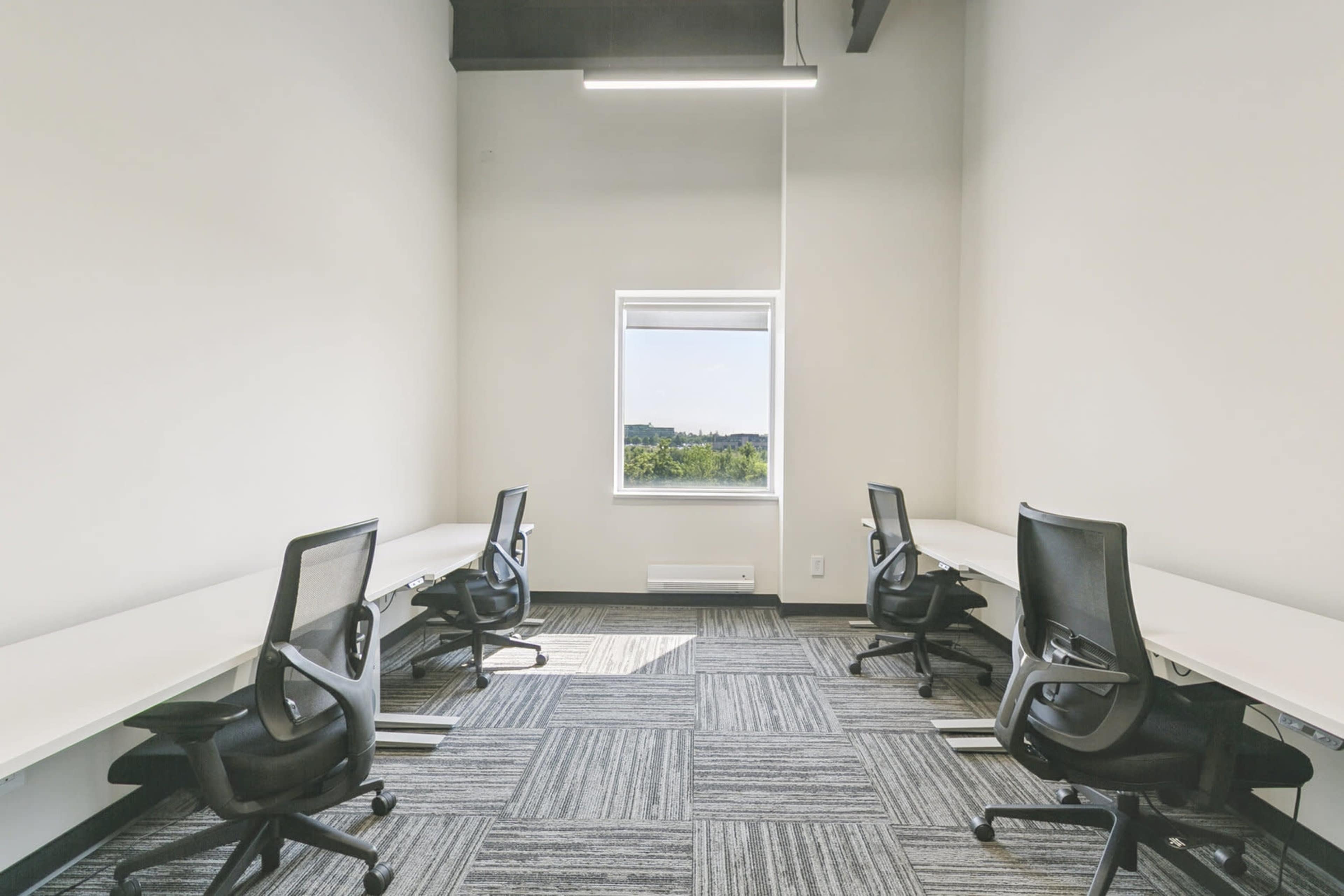 The image shows a small office space with four black office chairs and white desks arranged near a window, featuring gray carpet tiles.