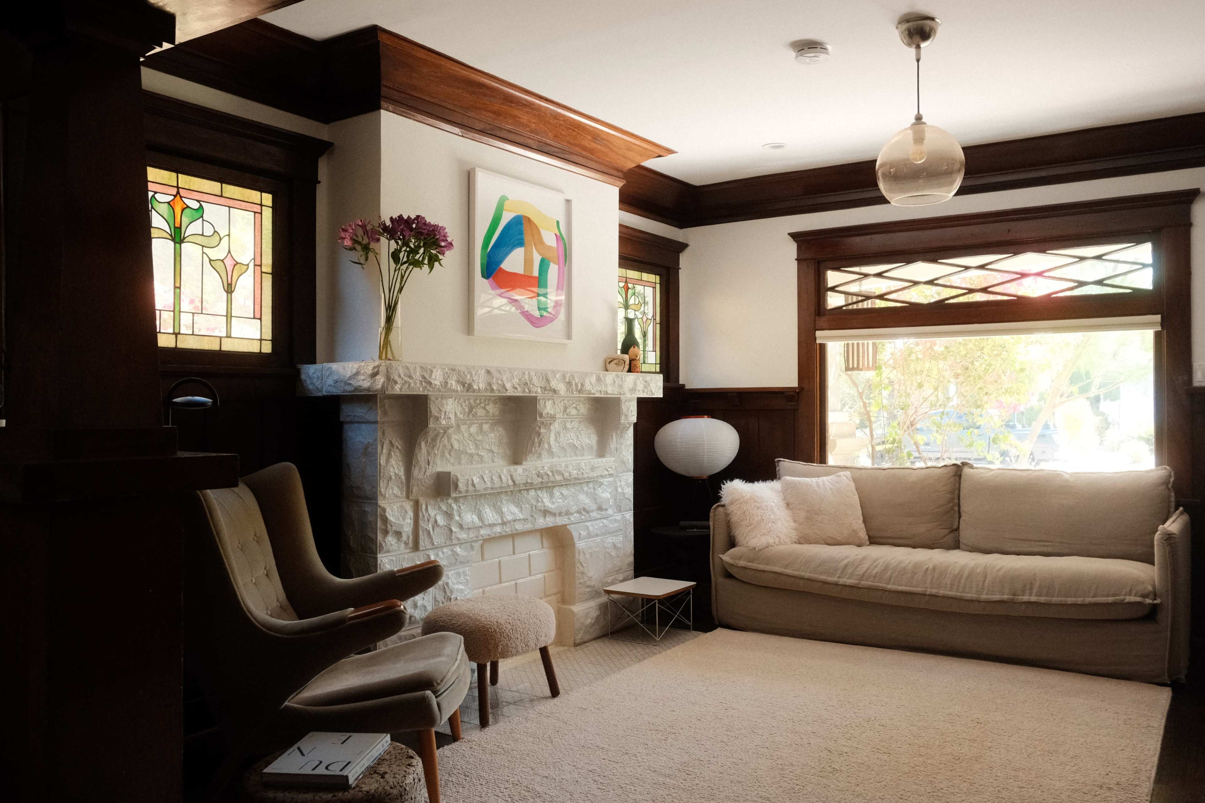 The image shows a living room with a light-colored sofa, a decorative stone fireplace, and stained glass windows, complemented by wooden detailing and a light fixture.
