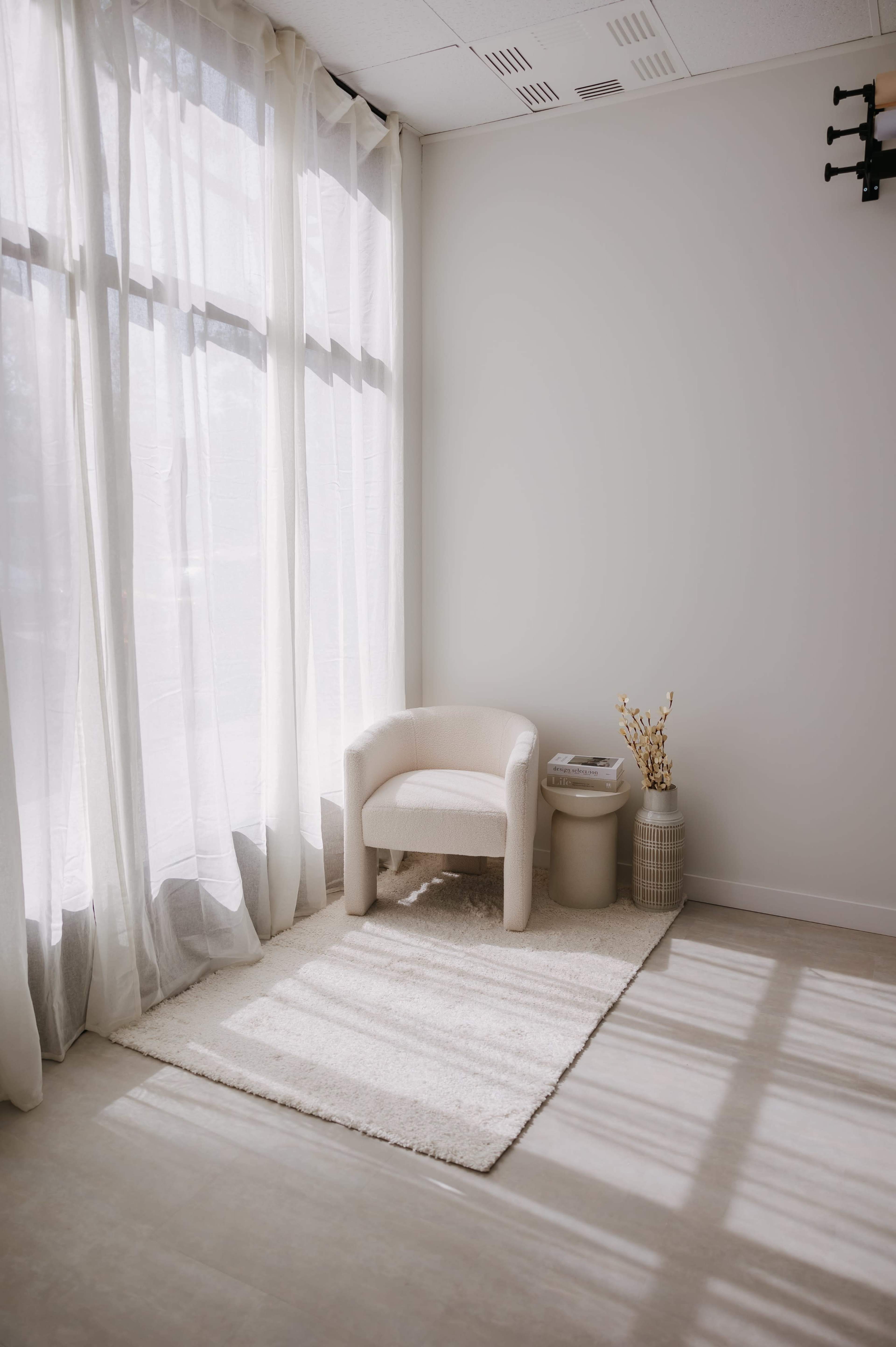 The image shows a minimalistic corner of a room featuring a light-colored armchair, a small side table, and a decorative vase beside a large window with sheer curtains.