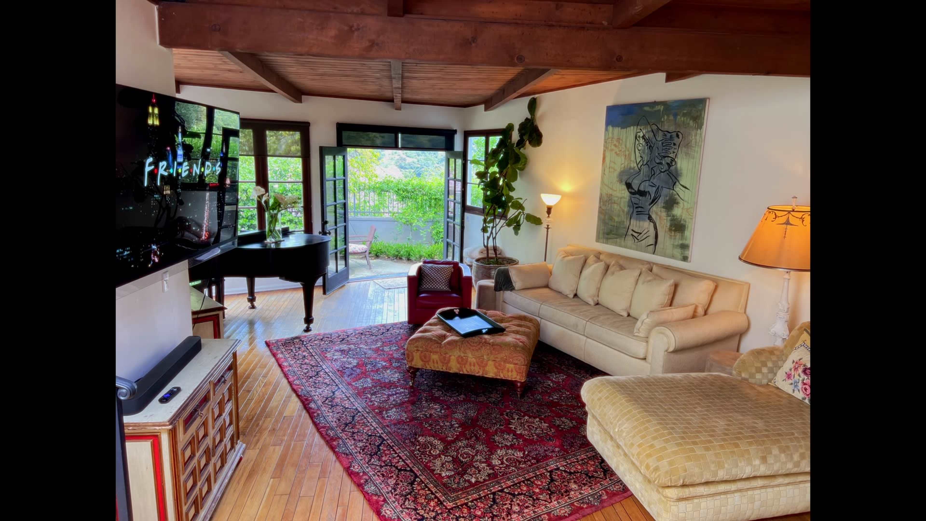 The image shows a living room with a sofa, an ottoman, a television displaying the show "Friends," a grand piano, and large windows overlooking a garden.