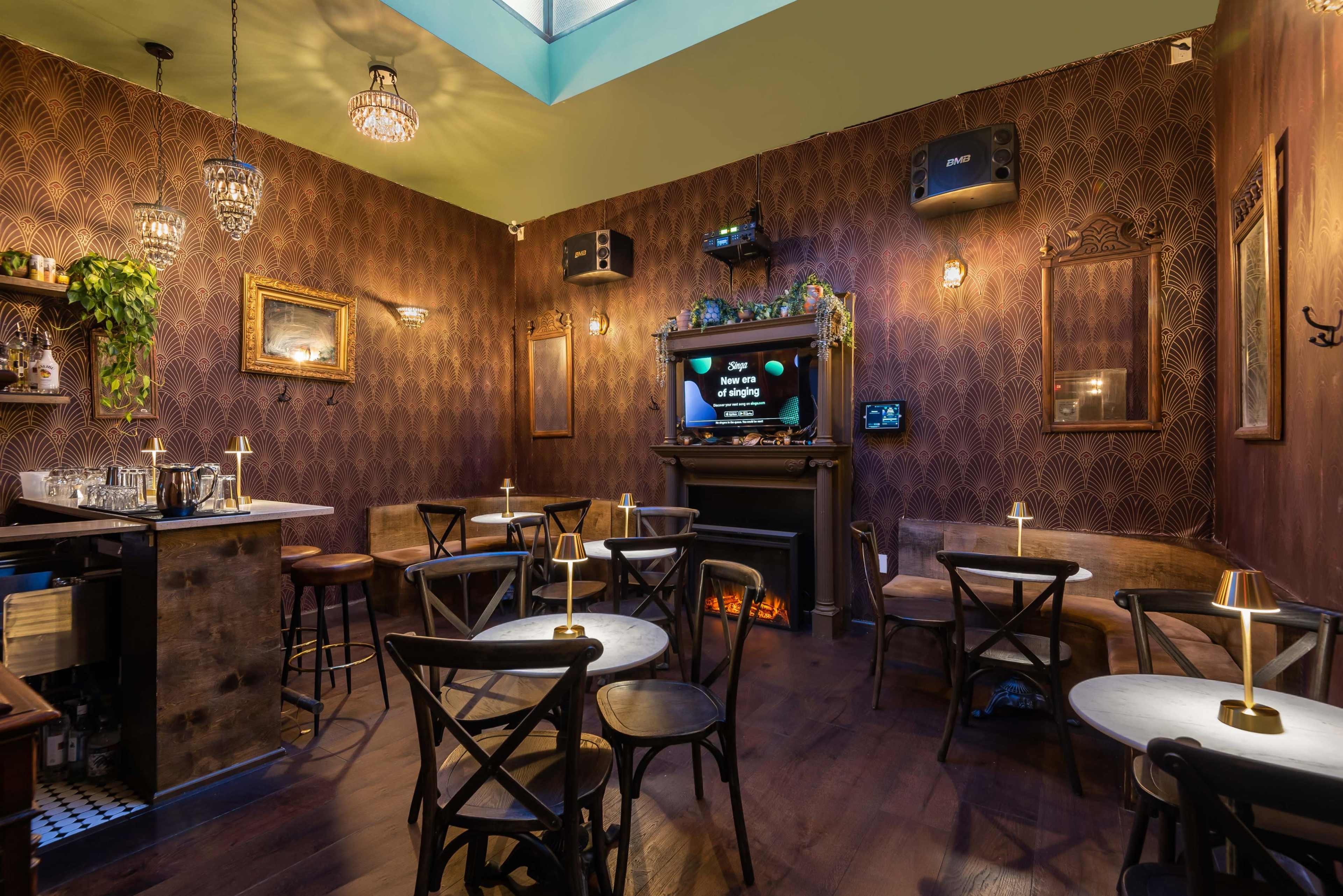 The image shows a cozy bar interior with wooden tables, a small bar area, and a wall-mounted TV, surrounded by vintage decor and warm lighting.