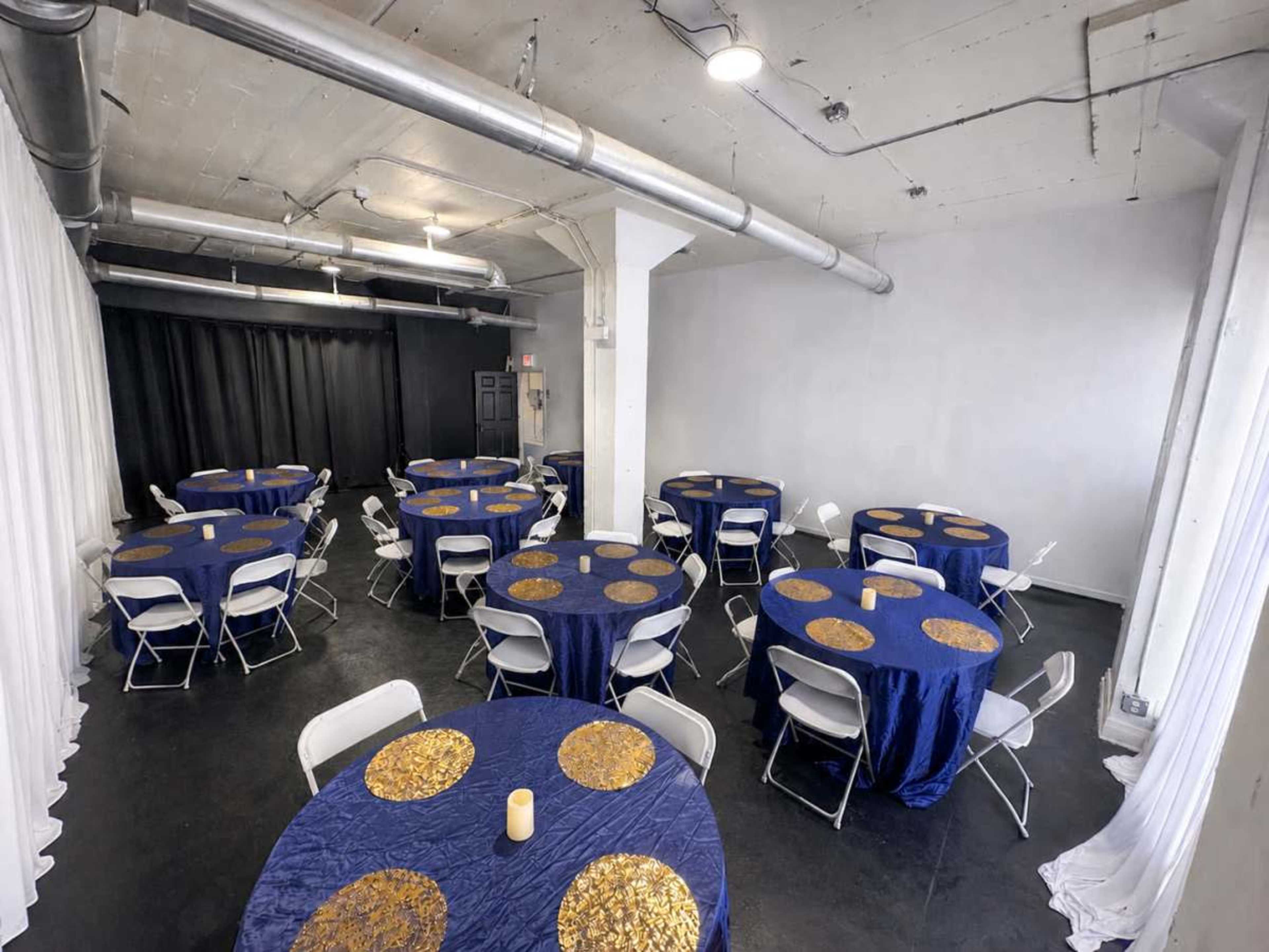 The image shows a spacious room set up for an event, featuring multiple tables covered with blue tablecloths adorned with gold circles, along with white folding chairs arranged around them.