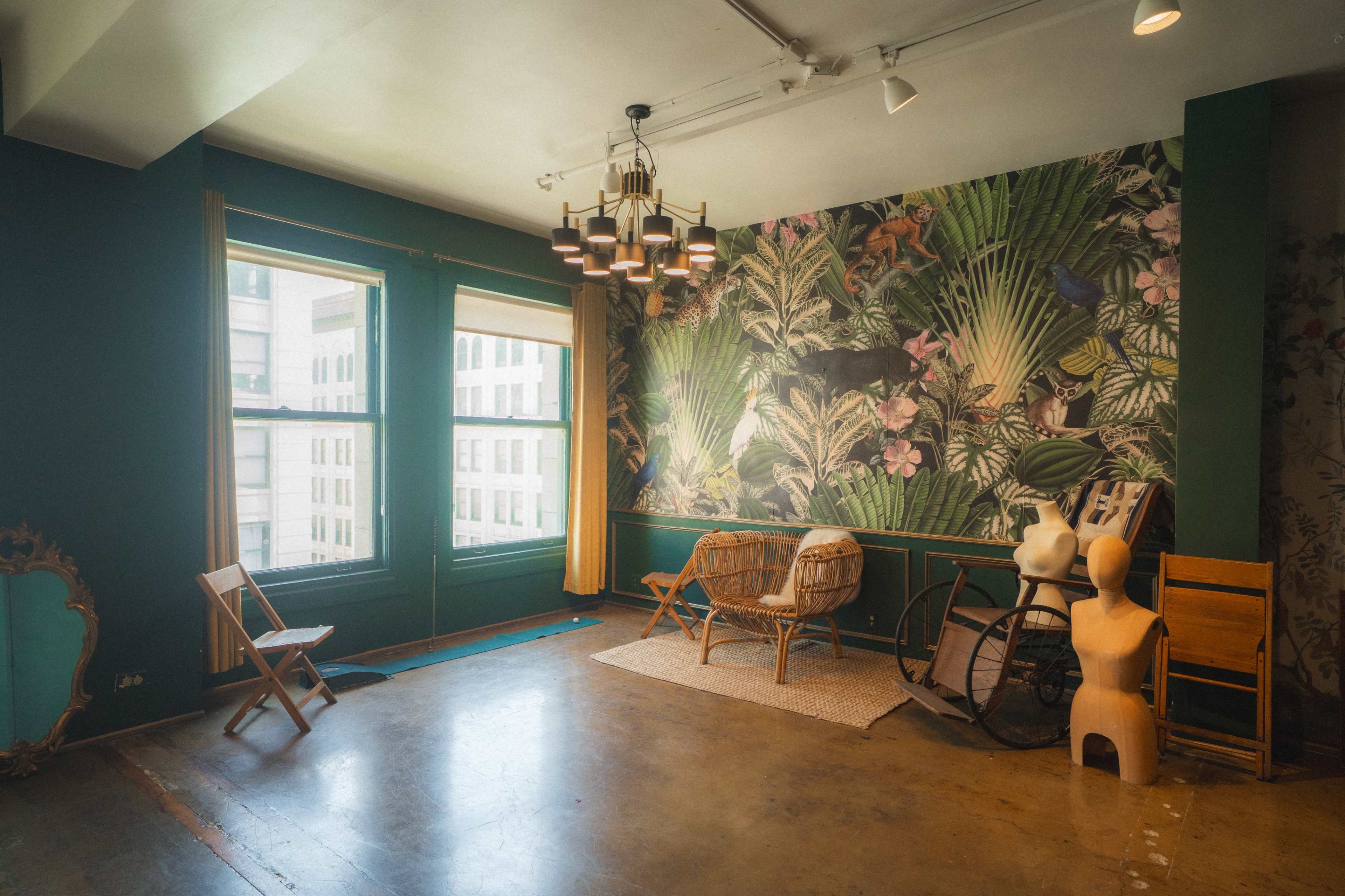 The image shows a room with a green accent wall featuring a large tropical mural, two large windows, and various pieces of furniture including wicker chairs and a wooden mannequin.