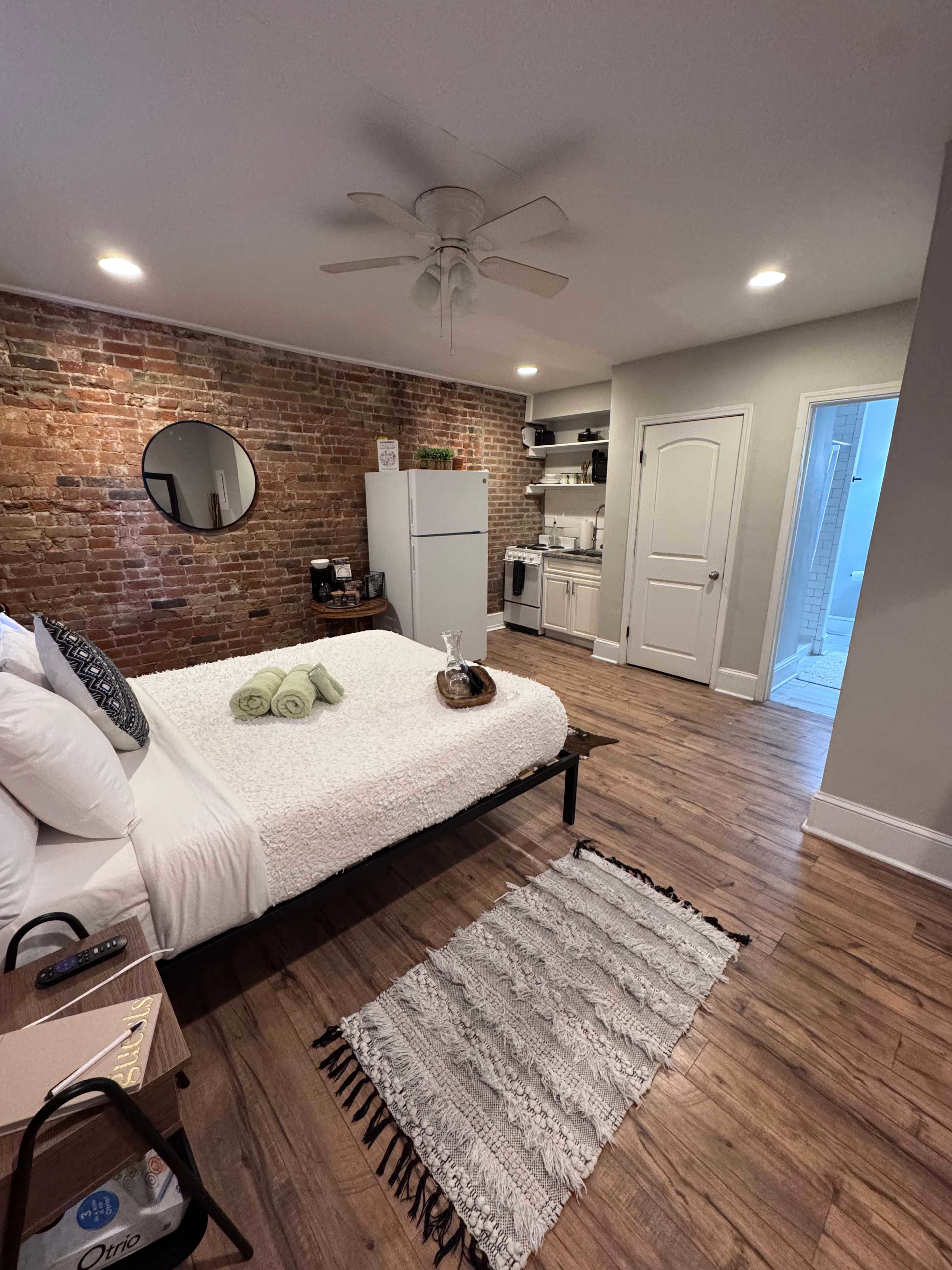 The image shows a cozy bedroom with a bed, a circular mirror on the wall, exposed brick, a kitchenette, and a small rug on a wooden floor.