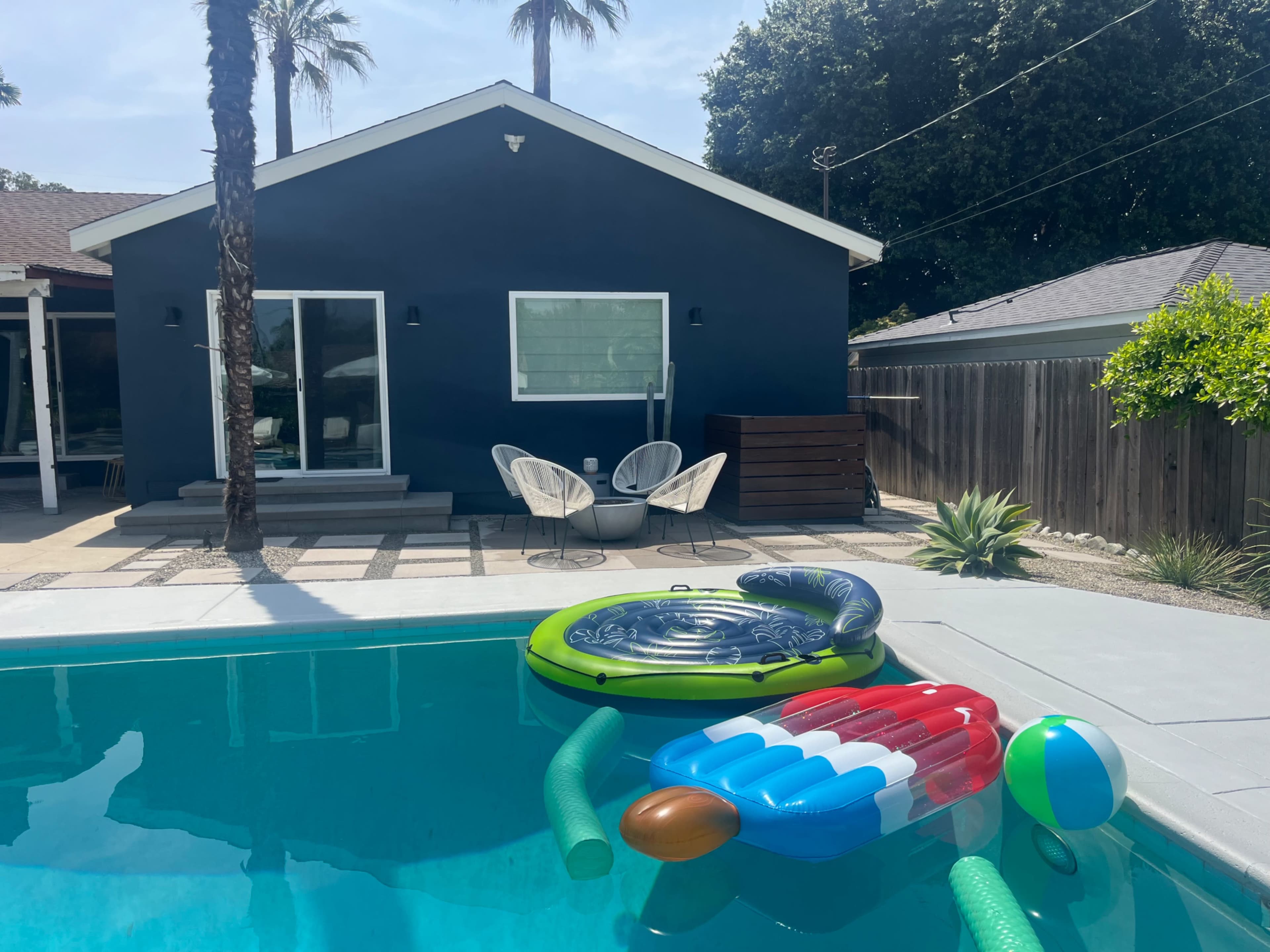 A swimming pool with colorful floats is in the foreground, while a modern house with a dark exterior and a patio is in the background, surrounded by palm trees.
