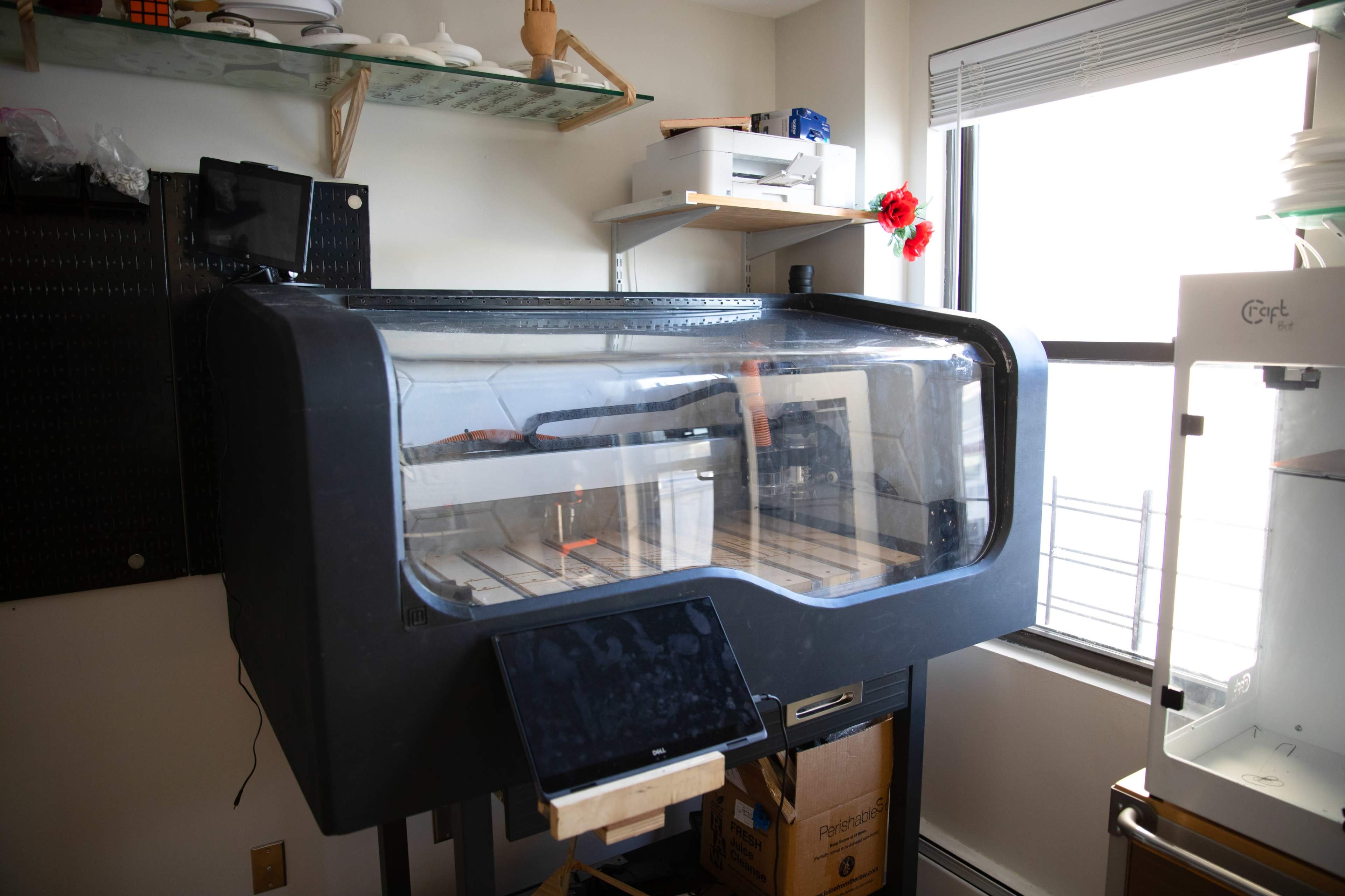 The image shows a large, enclosed laser cutter machine positioned in a well-lit workspace with shelves and tools in the background.