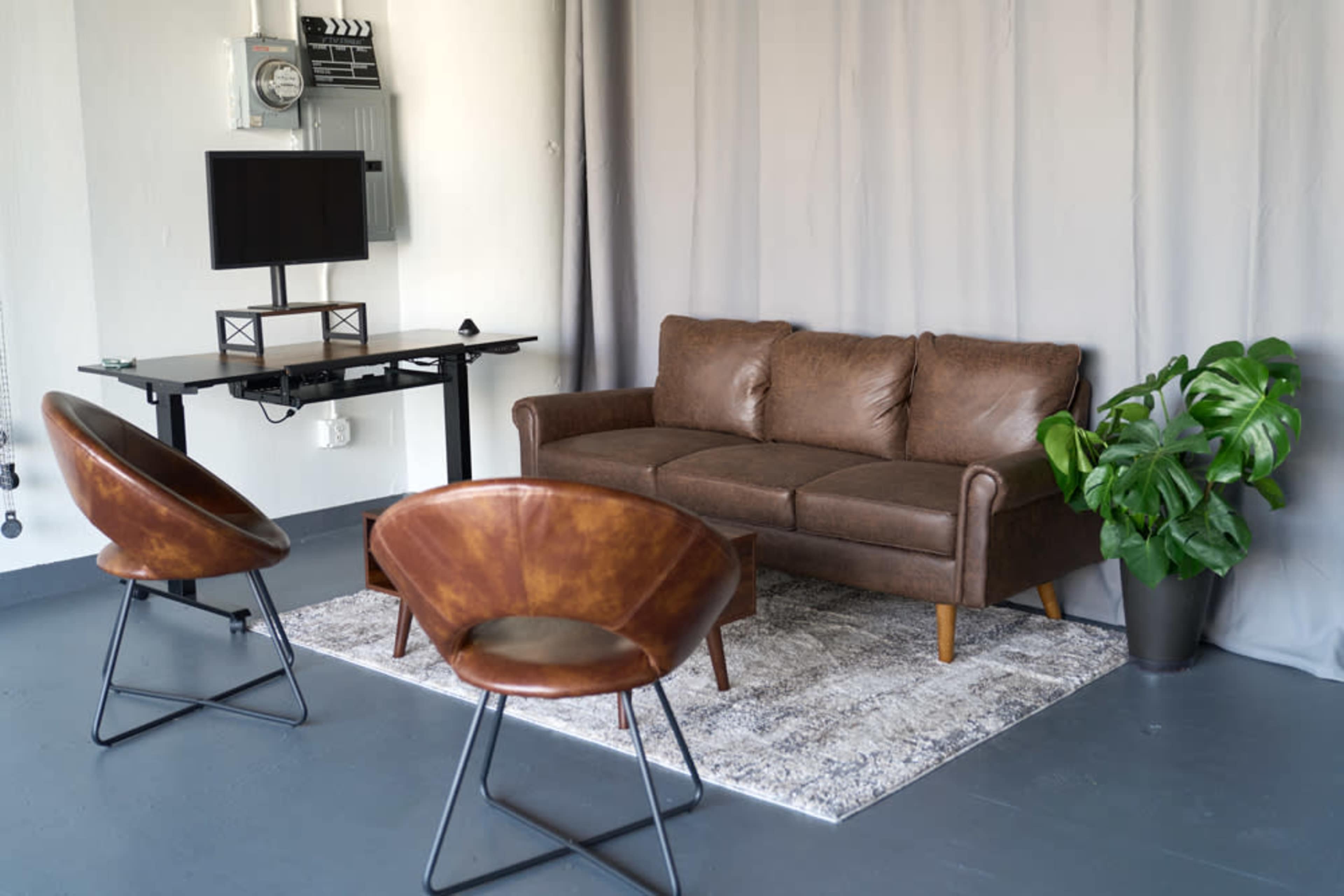 A minimalist interior with a brown leather sofa, two circular chairs, a small table, a TV on the wall, and a potted plant beside a gray curtain.
