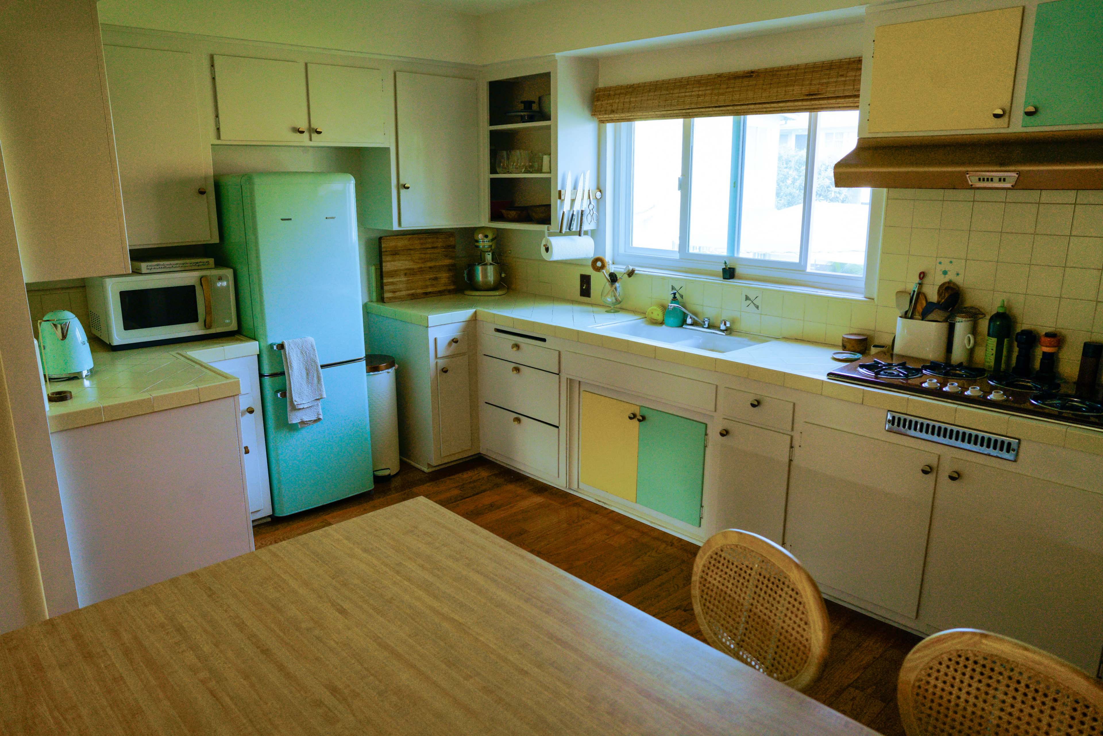 The image shows a brightly lit kitchen with mint green and yellow accents, featuring a vintage refrigerator, a stove, and a wooden dining table.