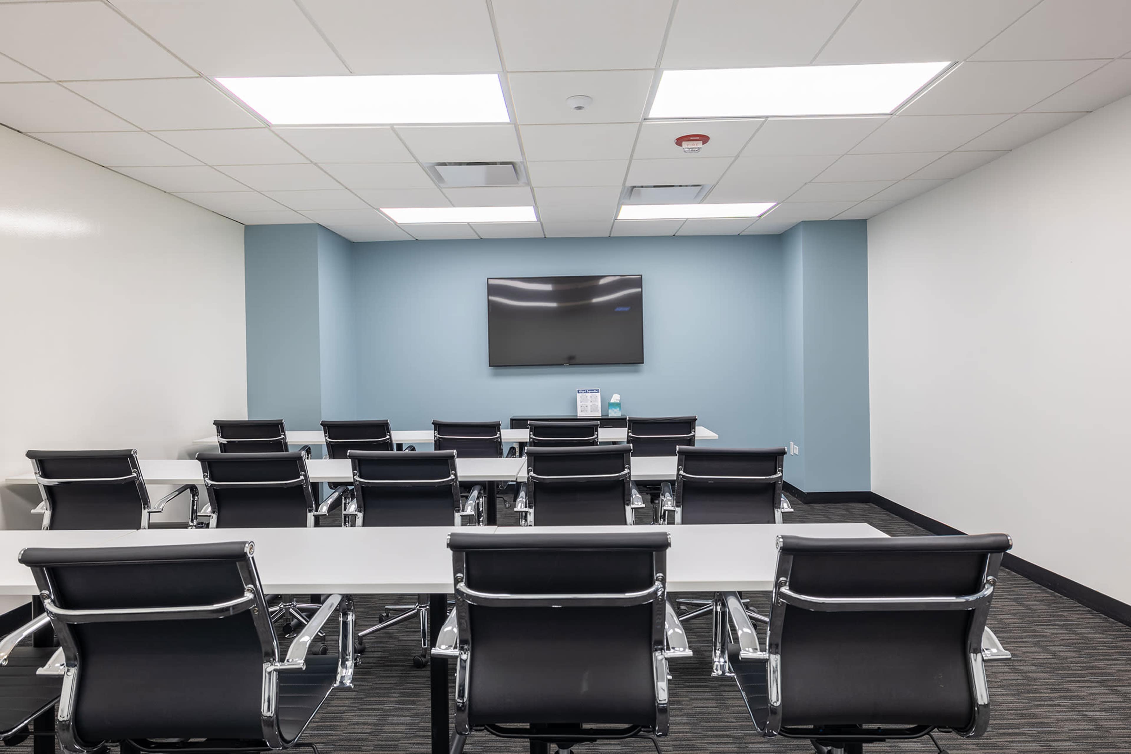 The image shows a modern conference room with a large wall-mounted TV, a long table, and several black chairs arranged in rows.
