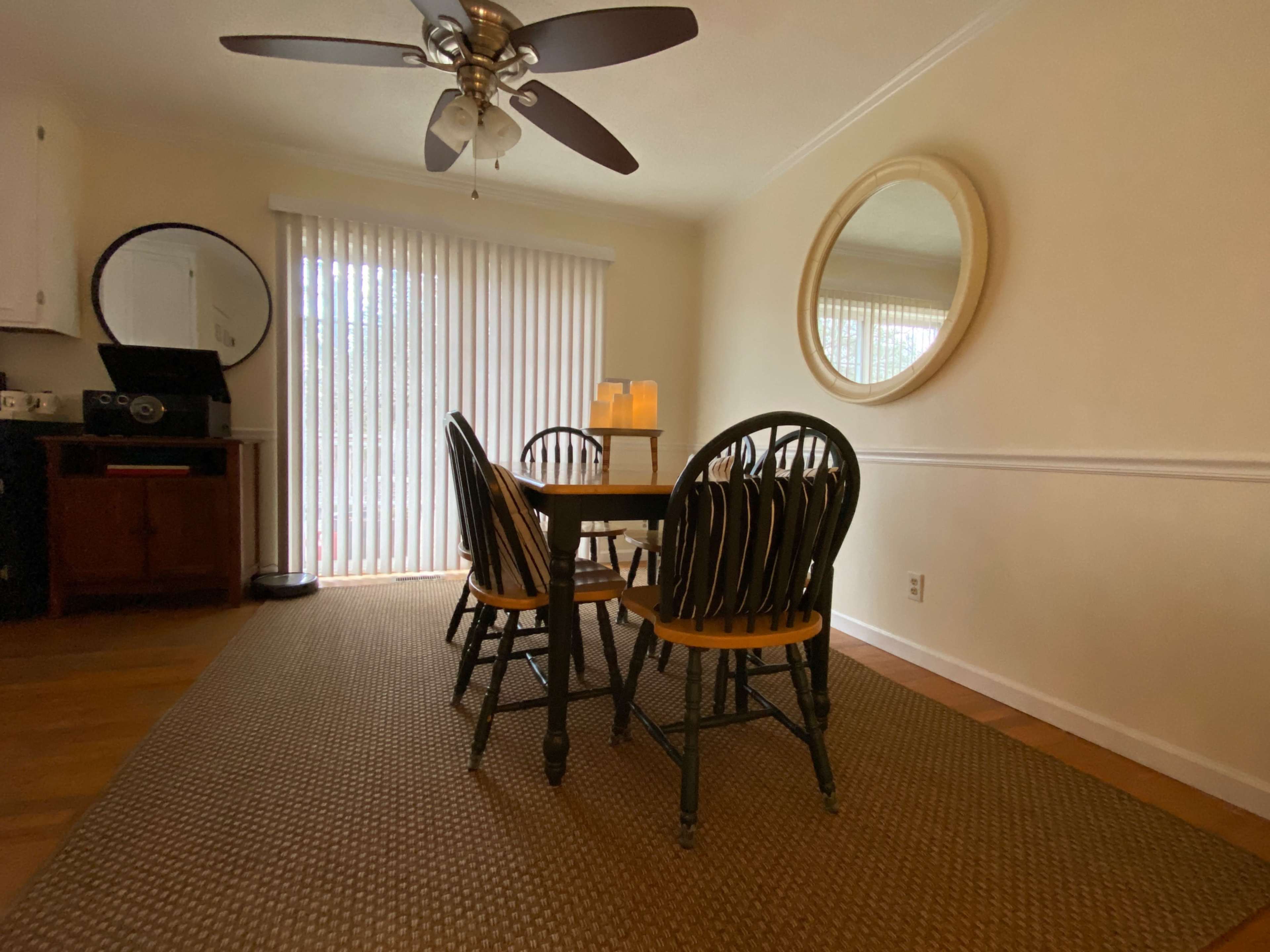 A dining room features a wooden table and four chairs, a ceiling fan, and a large mirror on the wall, with a sliding glass door covered by vertical blinds.
