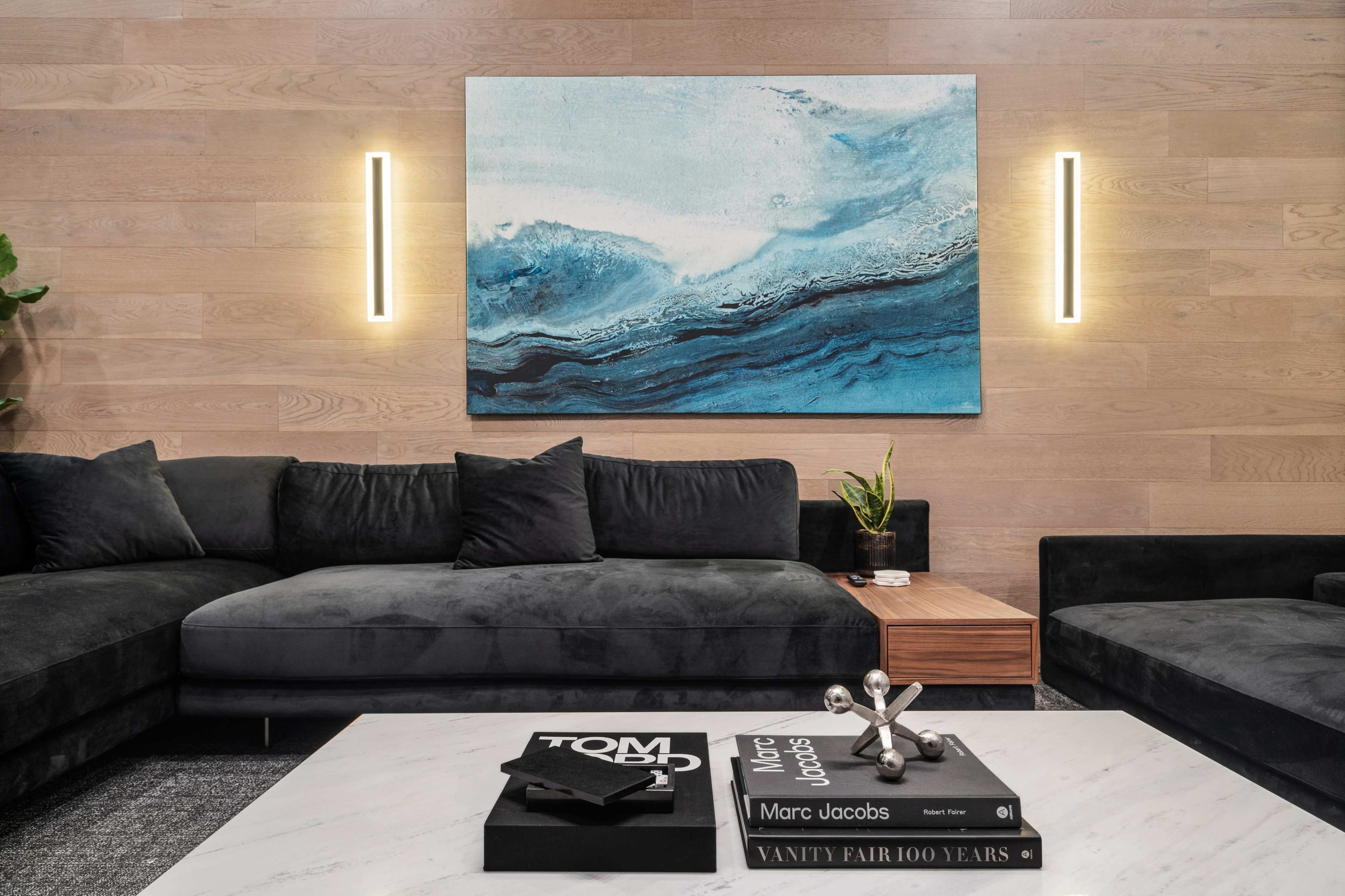 A modern living room features a dark sectional sofa, a wooden coffee table with stacked books, and a large abstract blue painting on the wall, illuminated by two wall sconces.