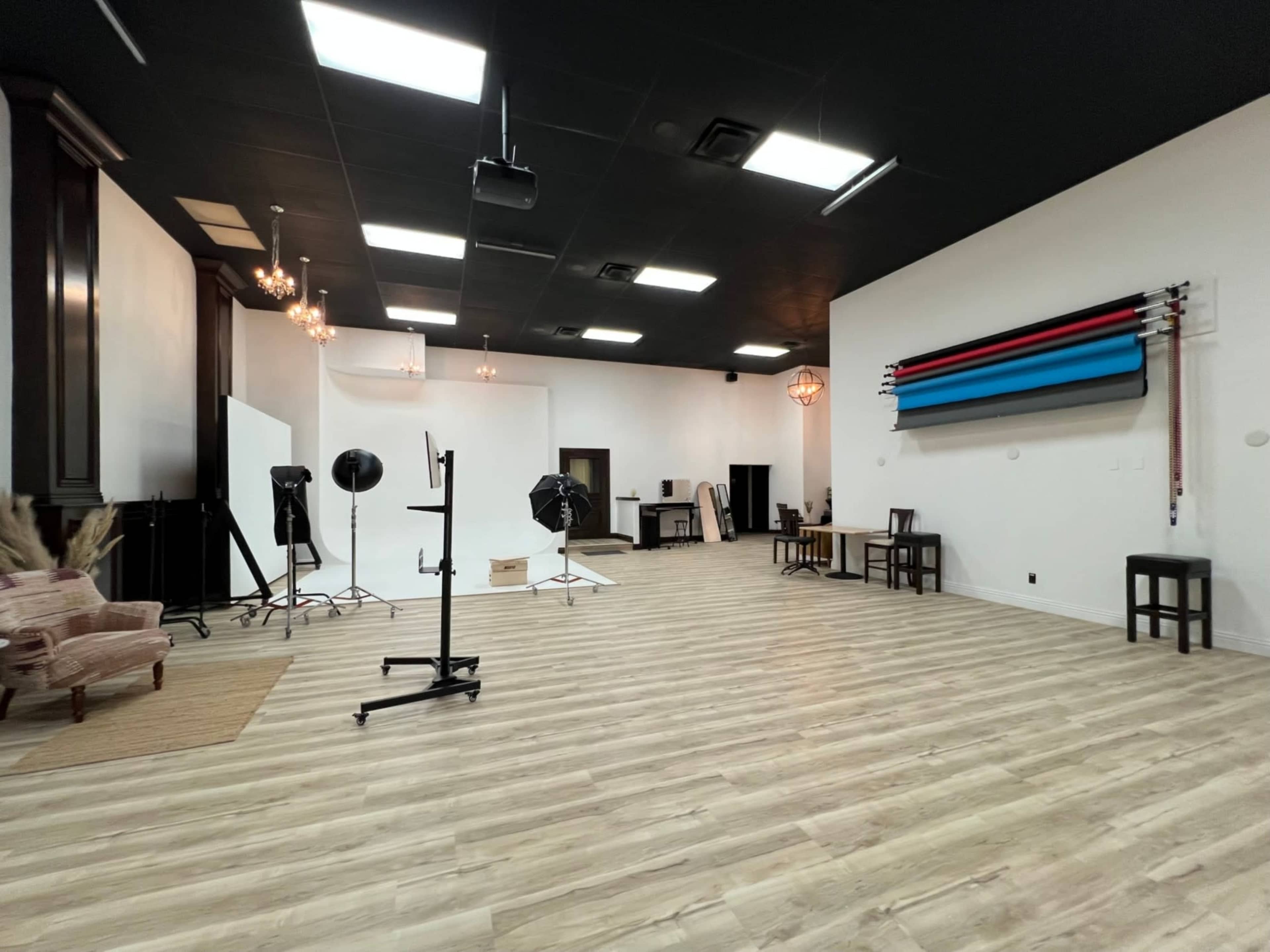 The image shows a spacious photography studio with wooden flooring, various lighting equipment, and colorful backdrops hanging on the wall.