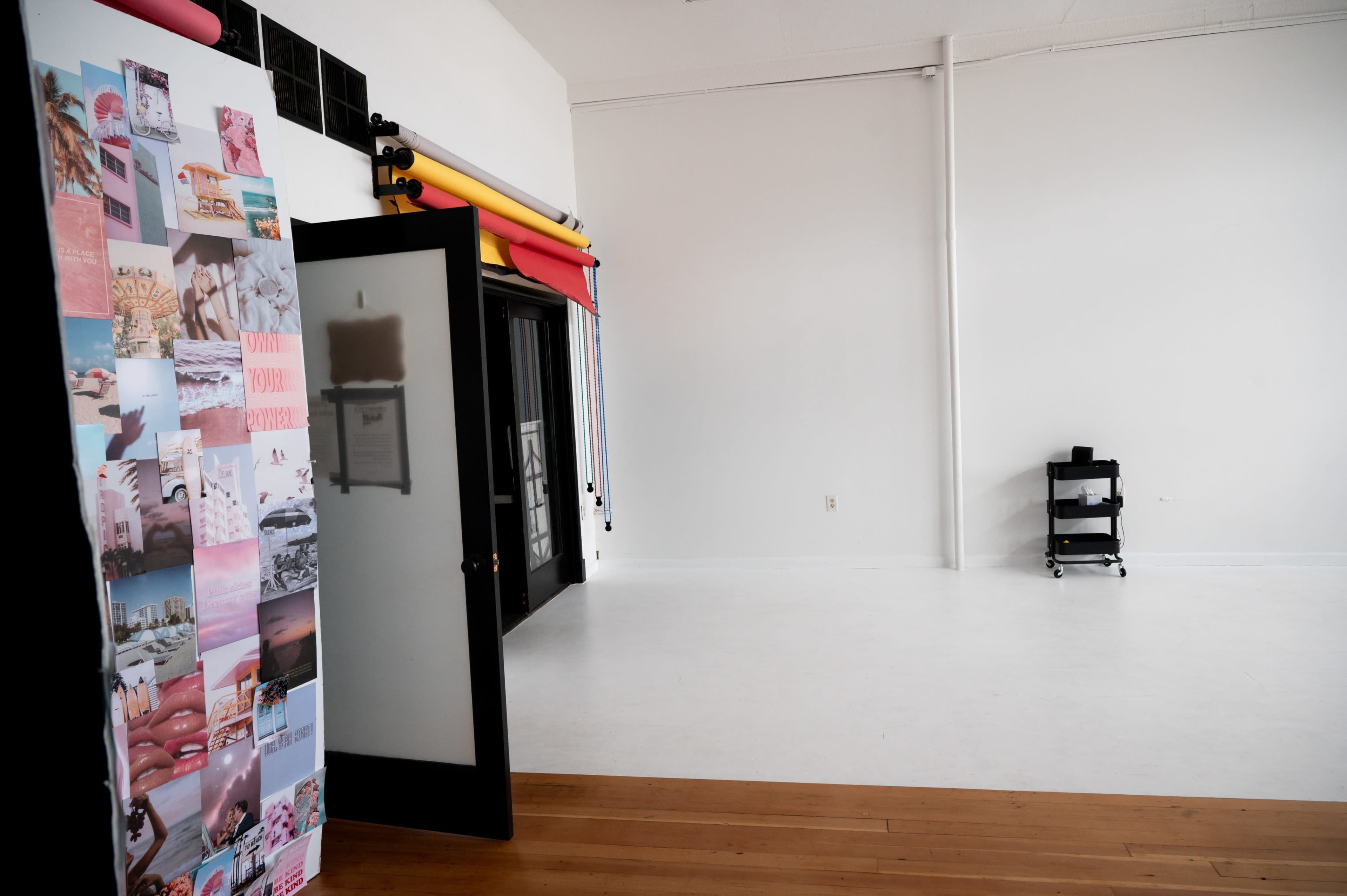 A spacious, brightly lit studio with a large wall decorated with a collage of photographs, and a door leading into the room that opens into a minimalist space featuring a black cart.