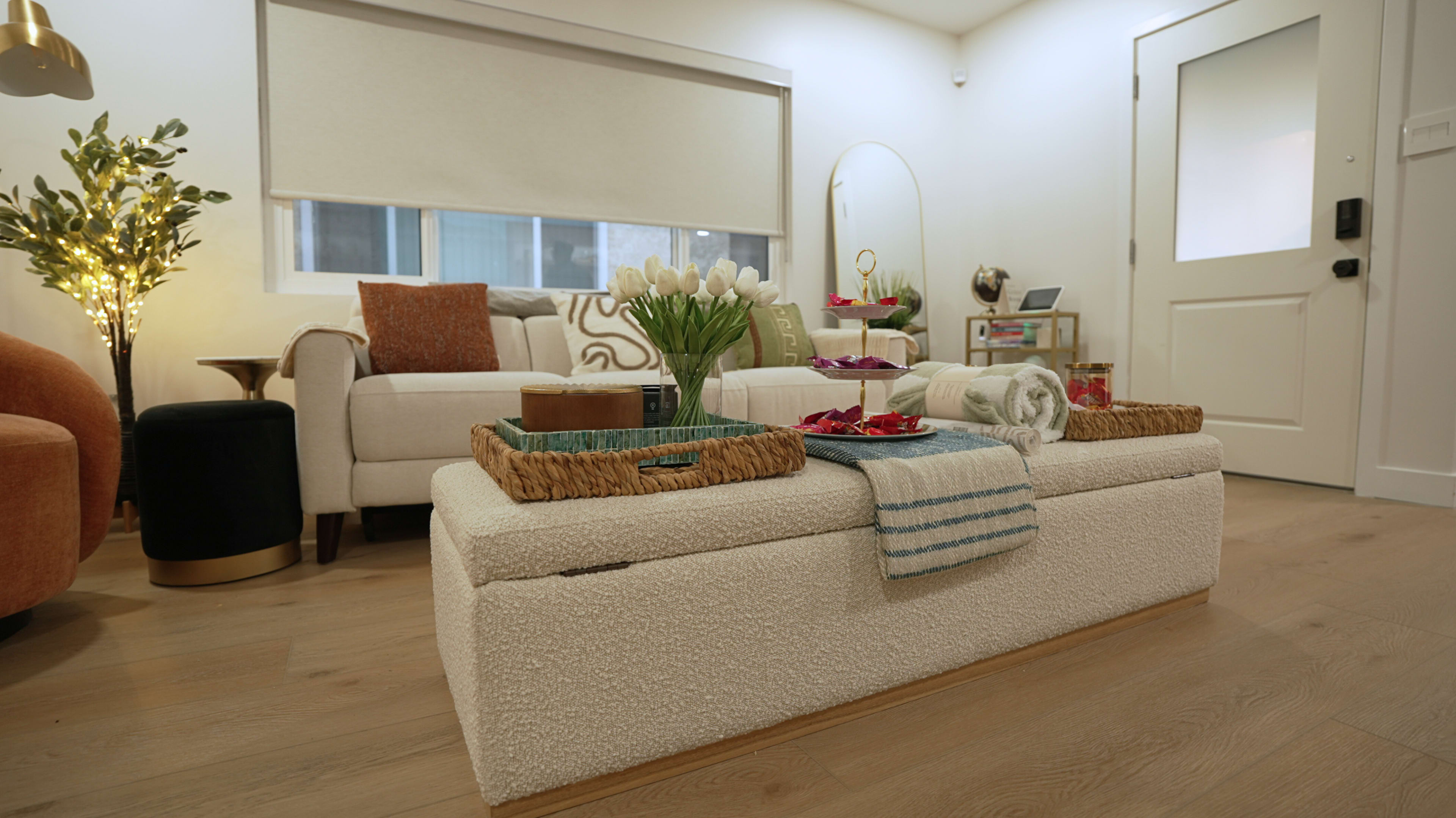 The image depicts a cozy living room featuring a light-colored sofa, a large ottoman with trays on top, and decorative items including flowers and towels, illuminated by soft lighting.