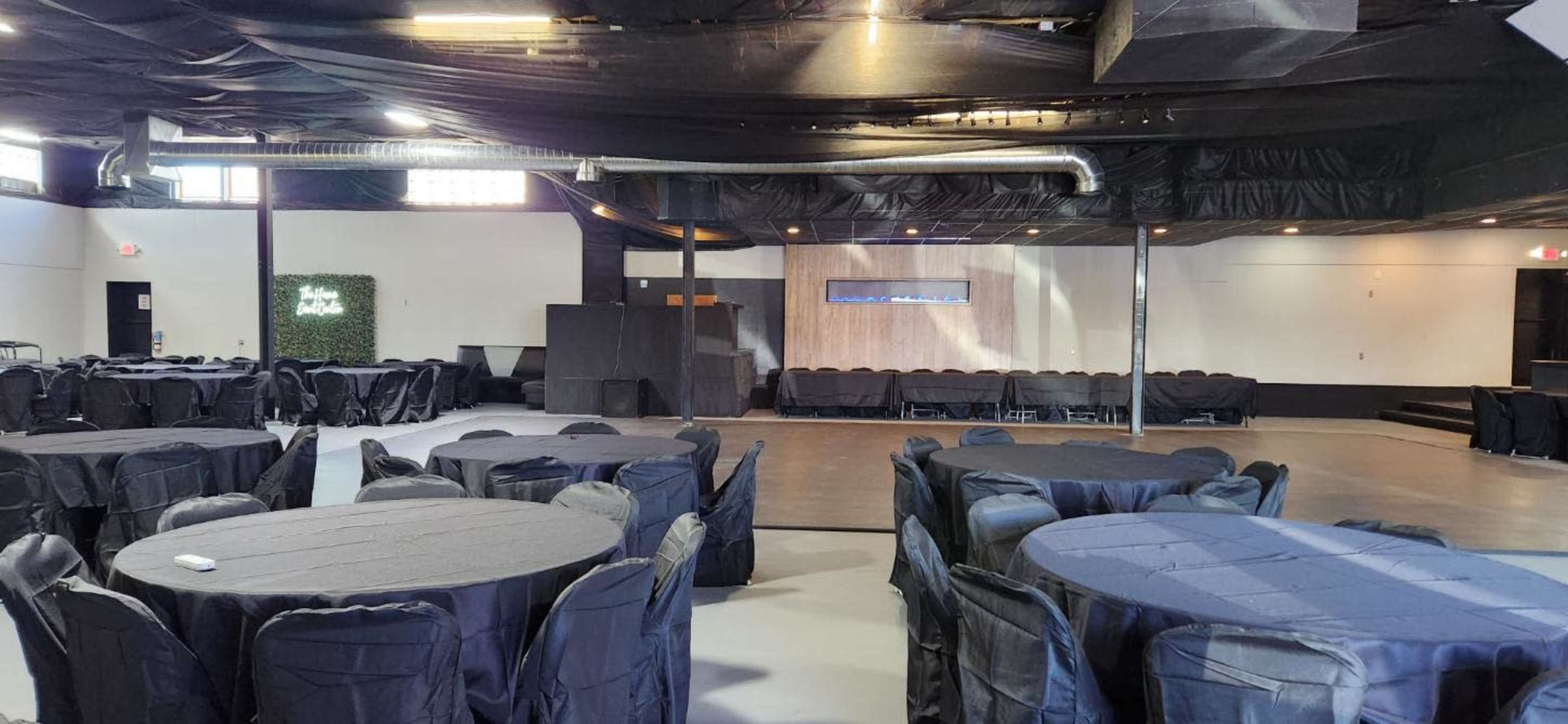 The image shows a large, empty indoor event space with round tables covered in black tablecloths and a small stage area at one end.