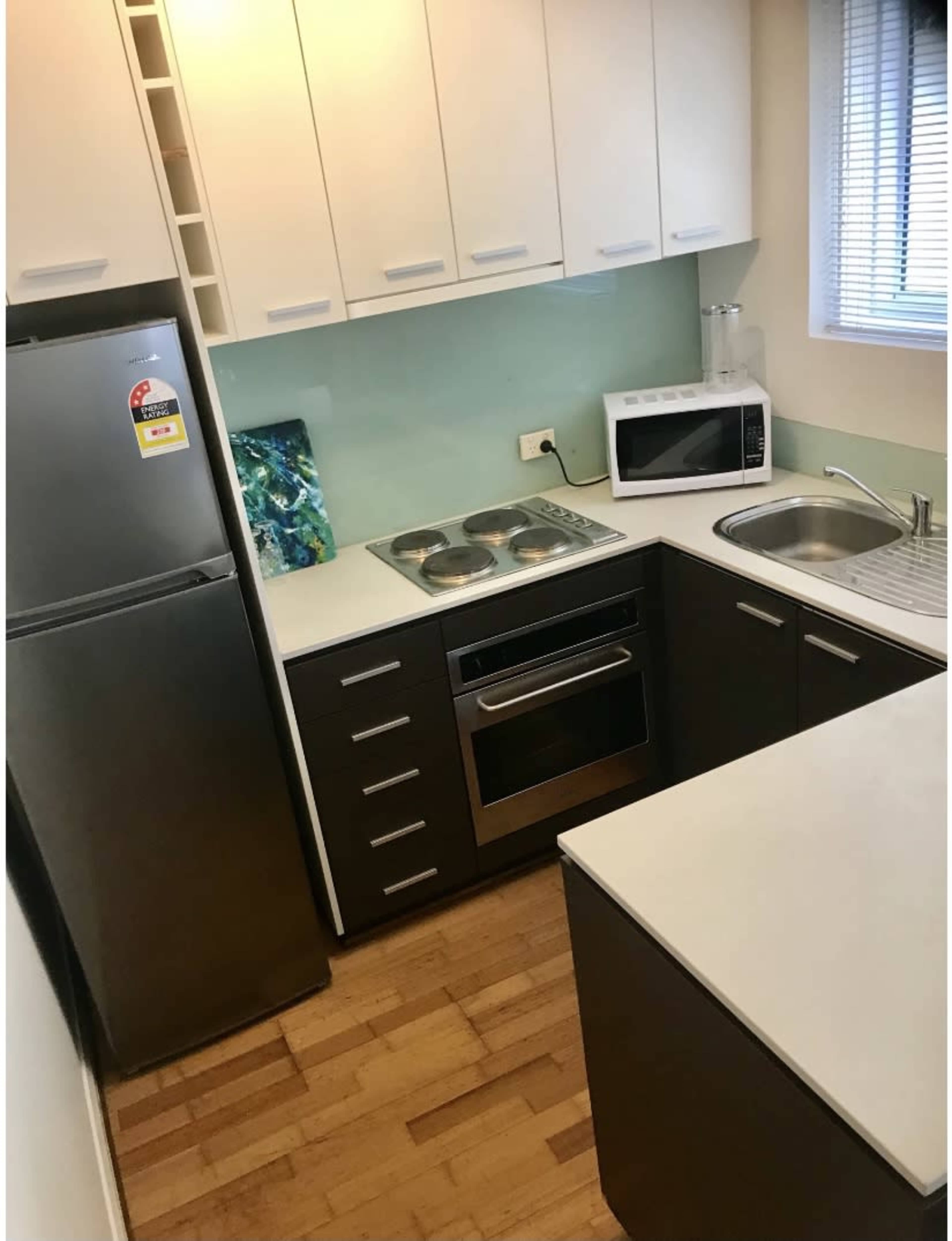 A small kitchen with a refrigerator, a sink, a stovetop, an oven, and a microwave, arranged along two walls with wooden flooring.