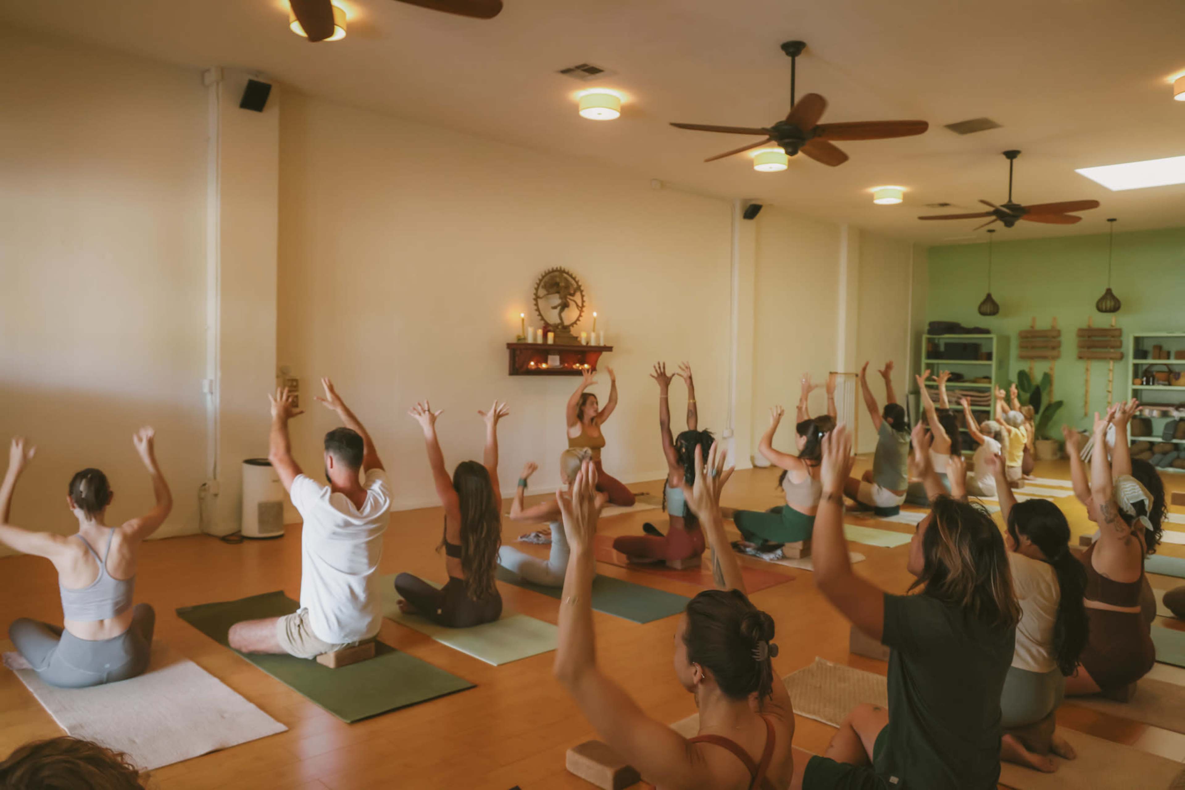 A group of individuals is practicing yoga in a spacious studio, with some seated on mats and others engaged in various poses while raising their arms.