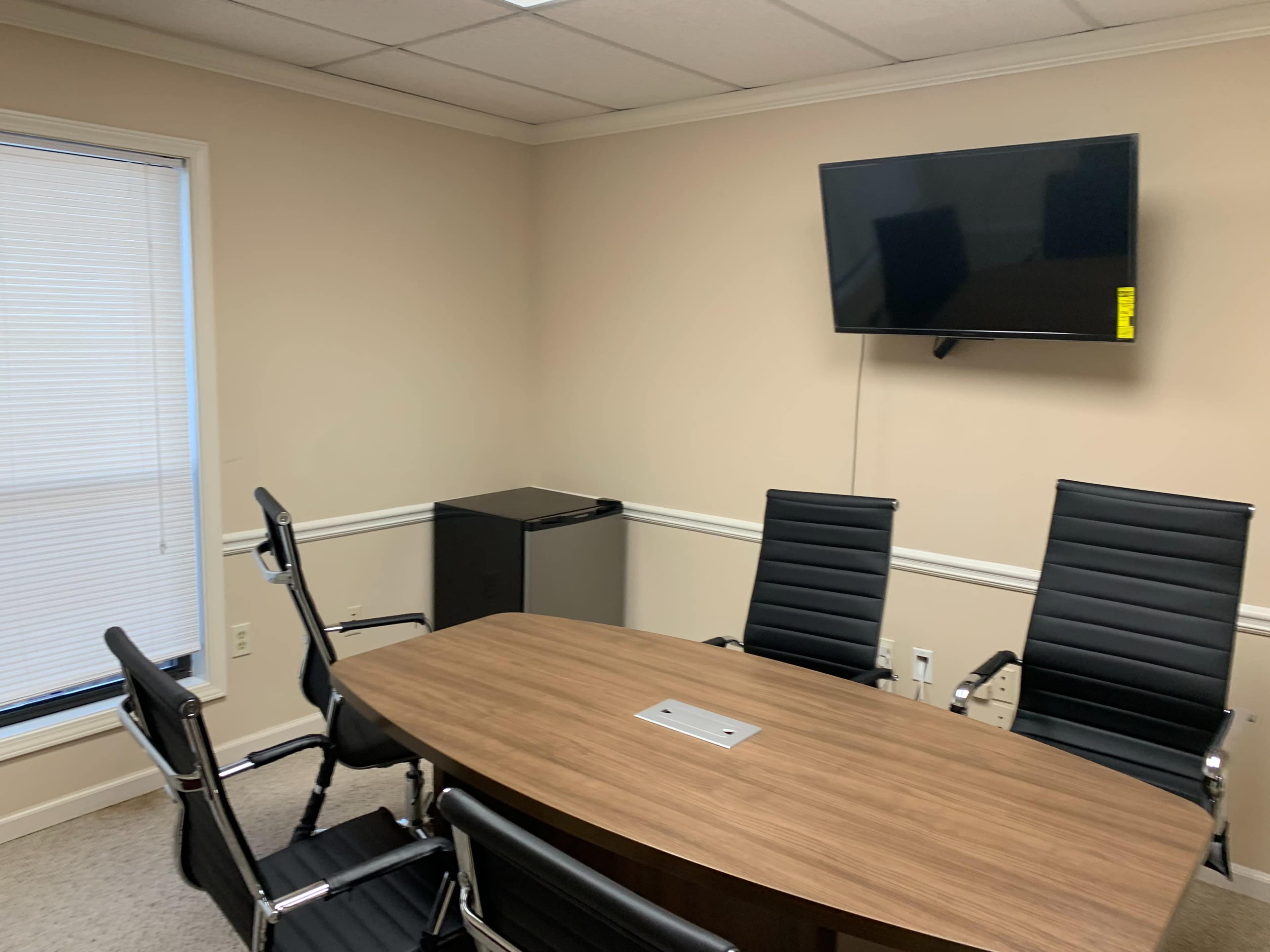 A conference room features a large wooden table, six black office chairs, a flat-screen TV mounted on the wall, and a small refrigerator in the corner.