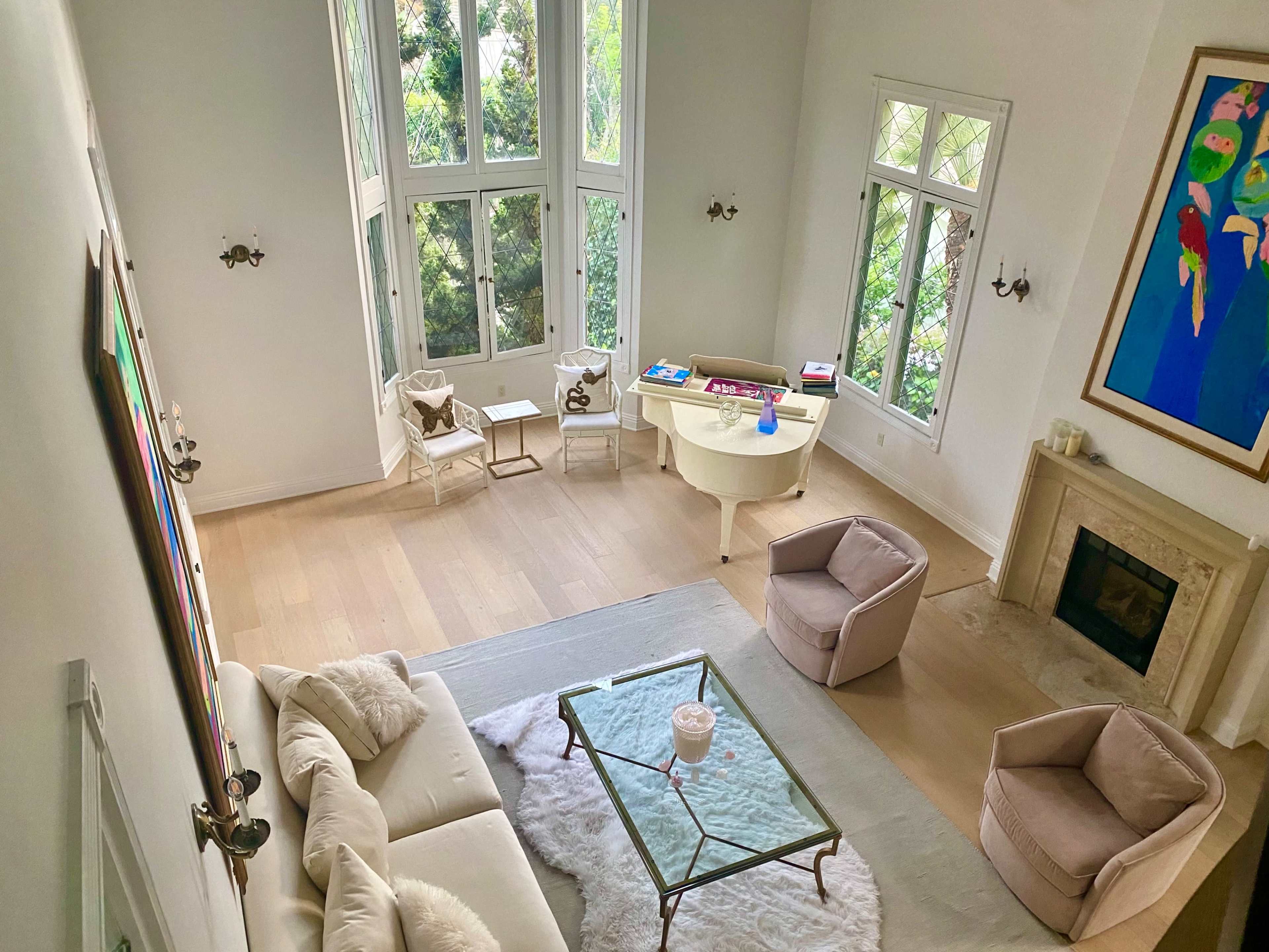 A spacious living room features large windows, a glass coffee table on a light rug, and soft seating arranged around the fireplace.