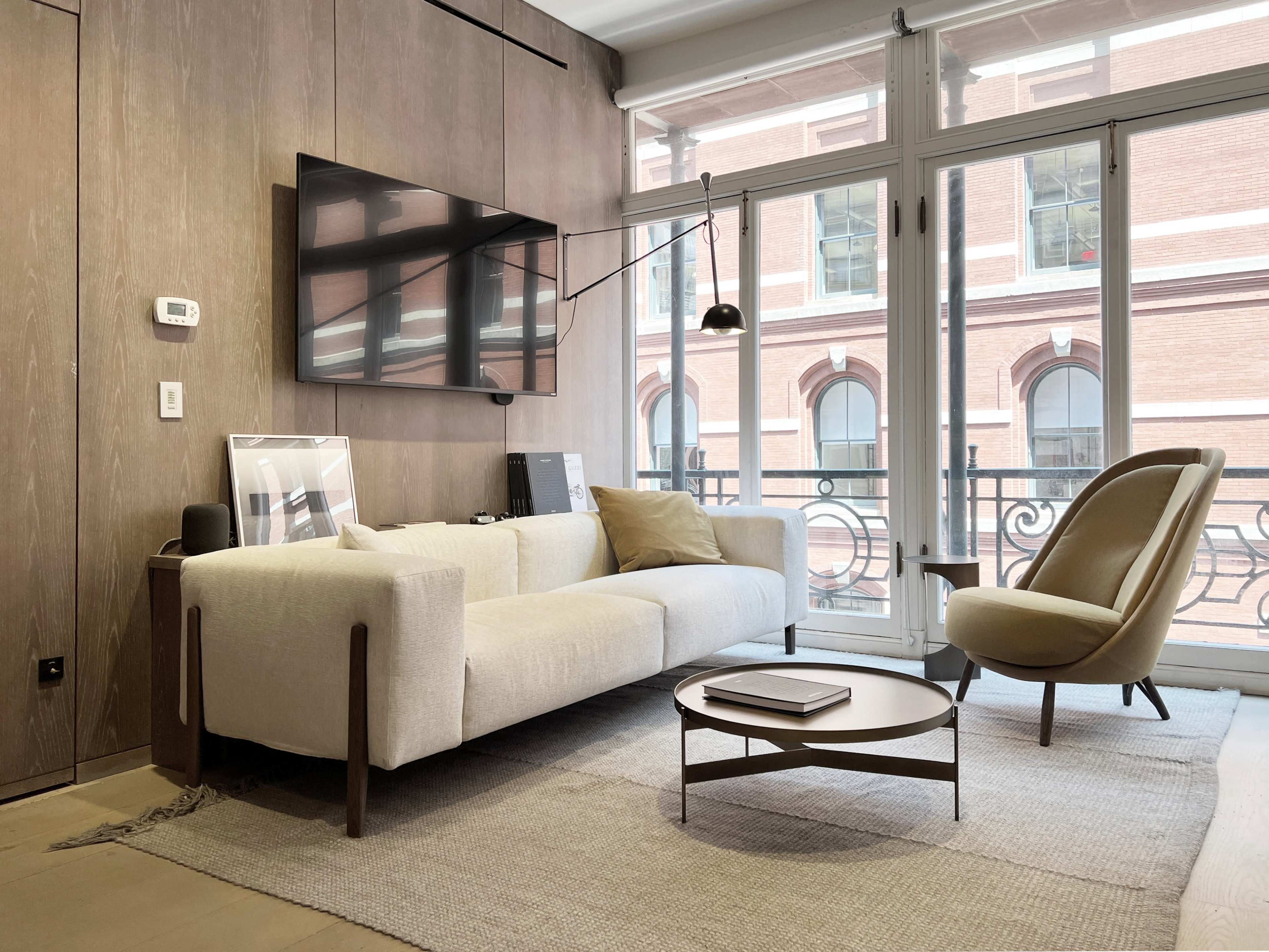 A modern living room features a light-colored sofa, a round coffee table, and a beige armchair, with large windows showcasing a view of a brick building outside.