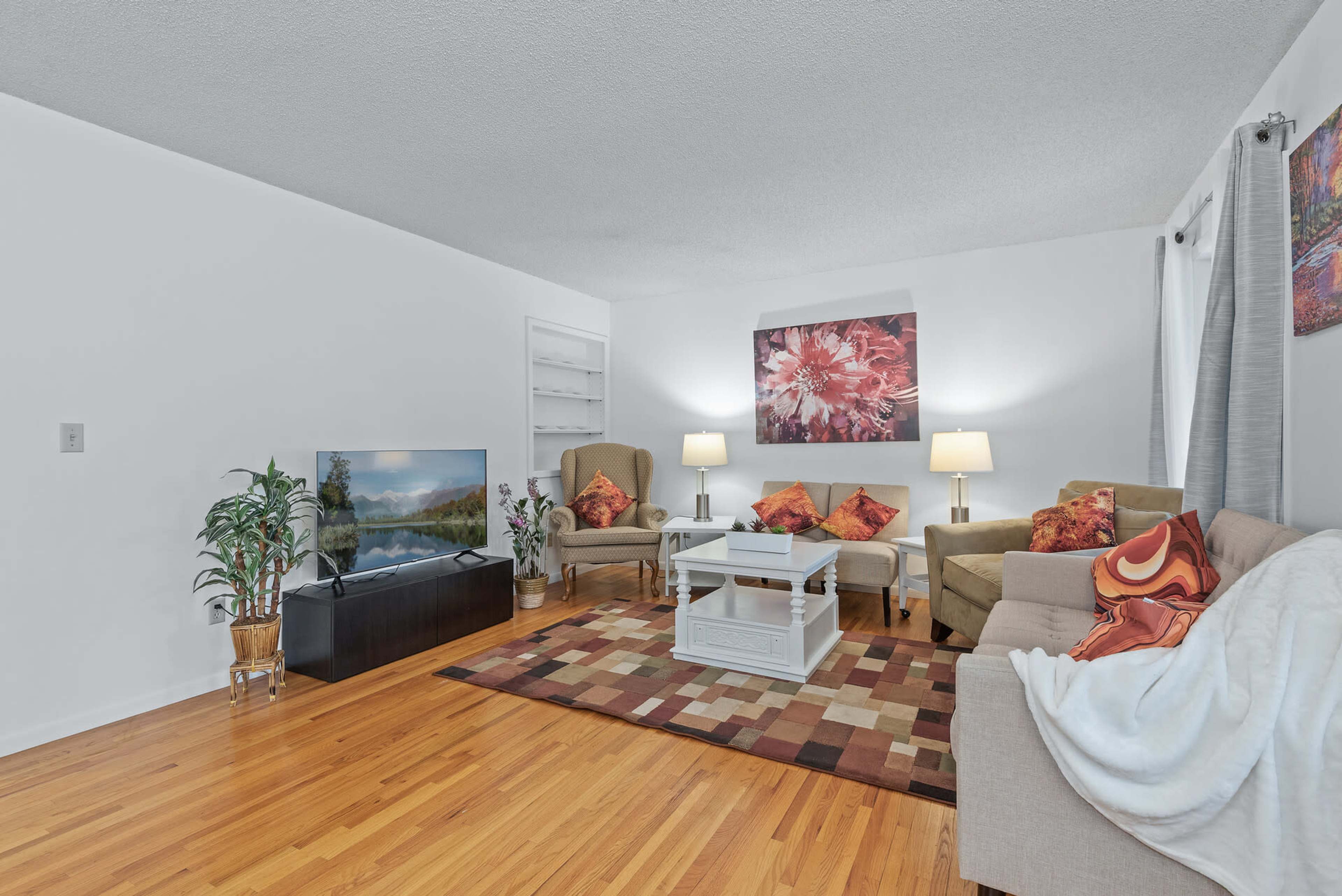 The living room features a beige sofa, two armchairs, a coffee table, and a television placed on a cabinet, all arranged around a colorful area rug.