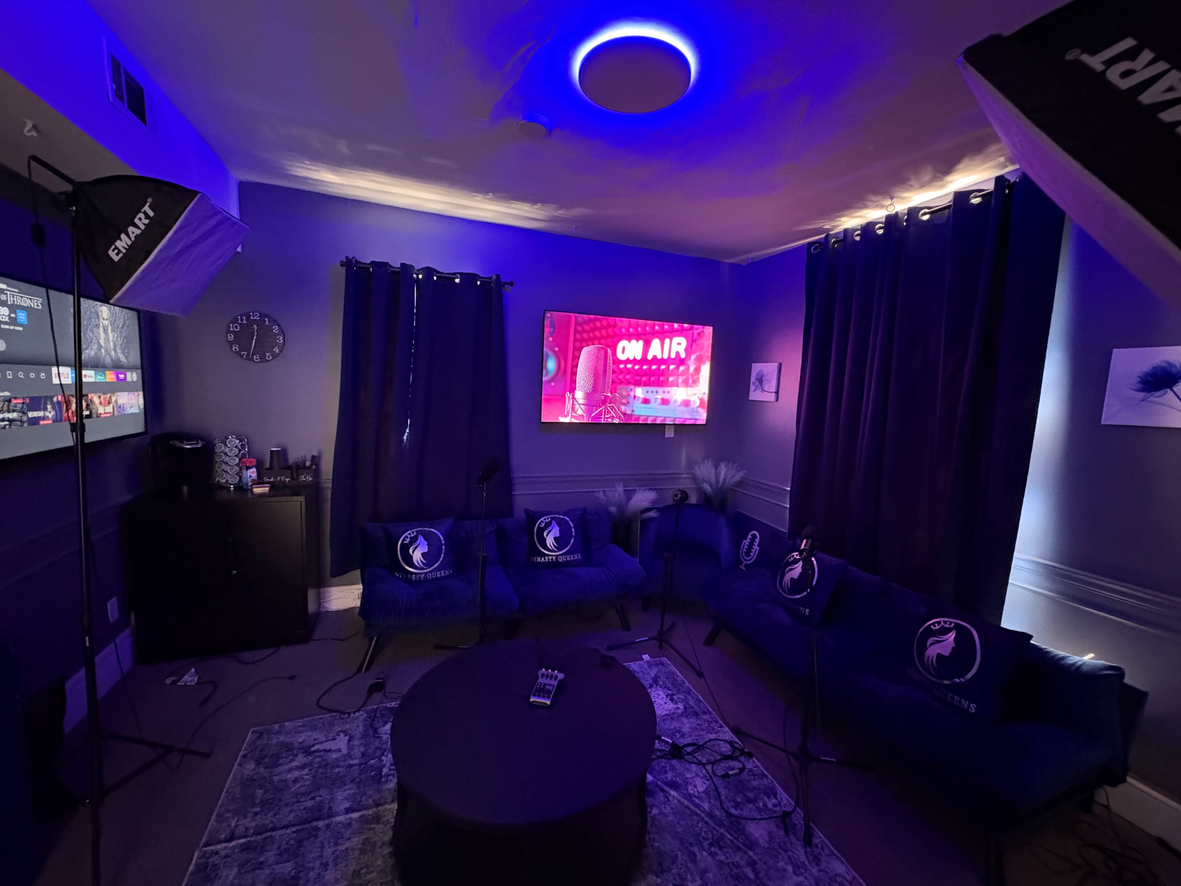 The image shows a modern recording studio with dark blue walls, a round table in the center, and sofas arranged for discussion, illuminated by colorful lights and featuring equipment for podcasting or streaming.