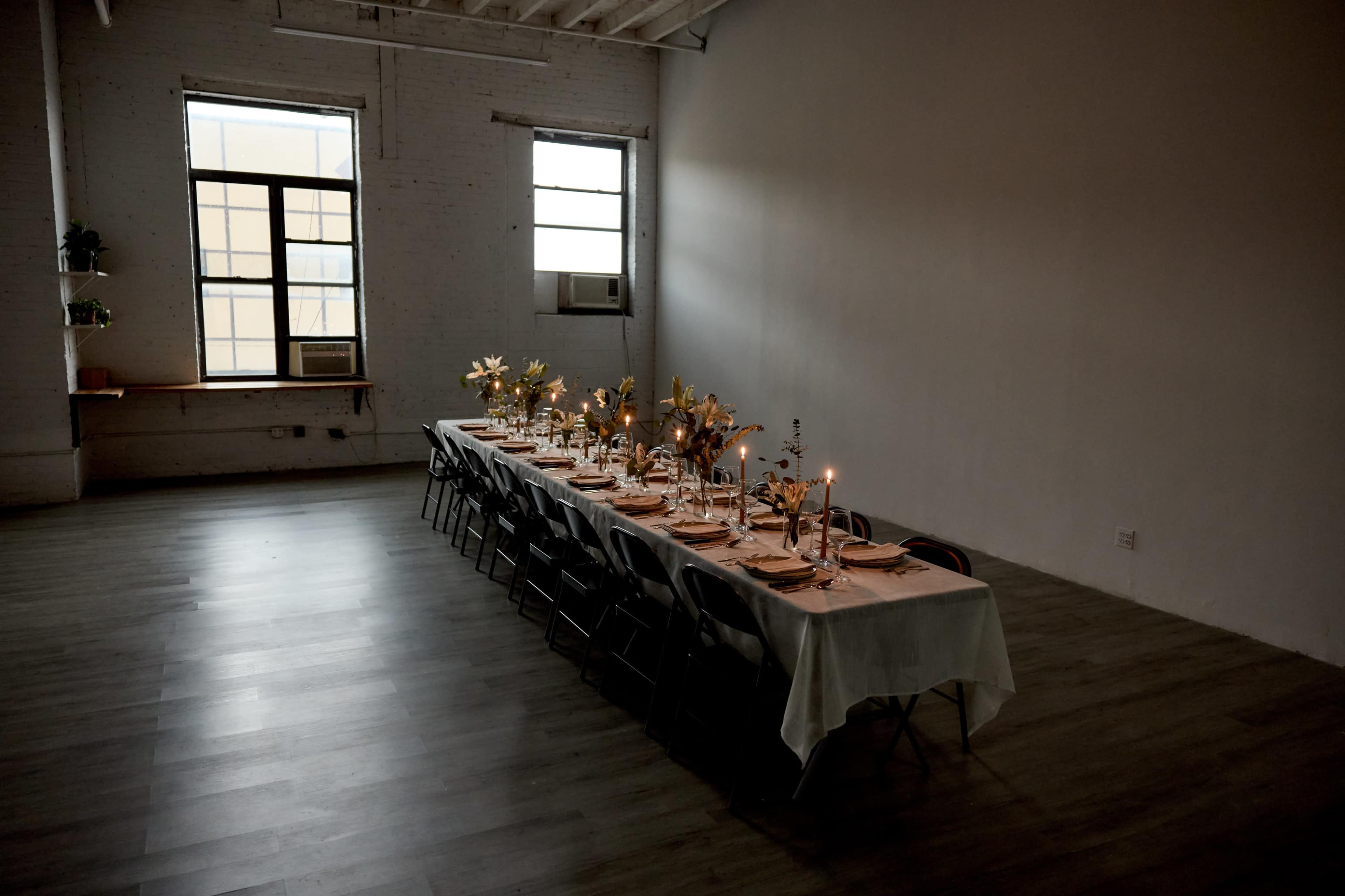 A long dining table is set for a meal in a sparsely decorated room with large windows.
