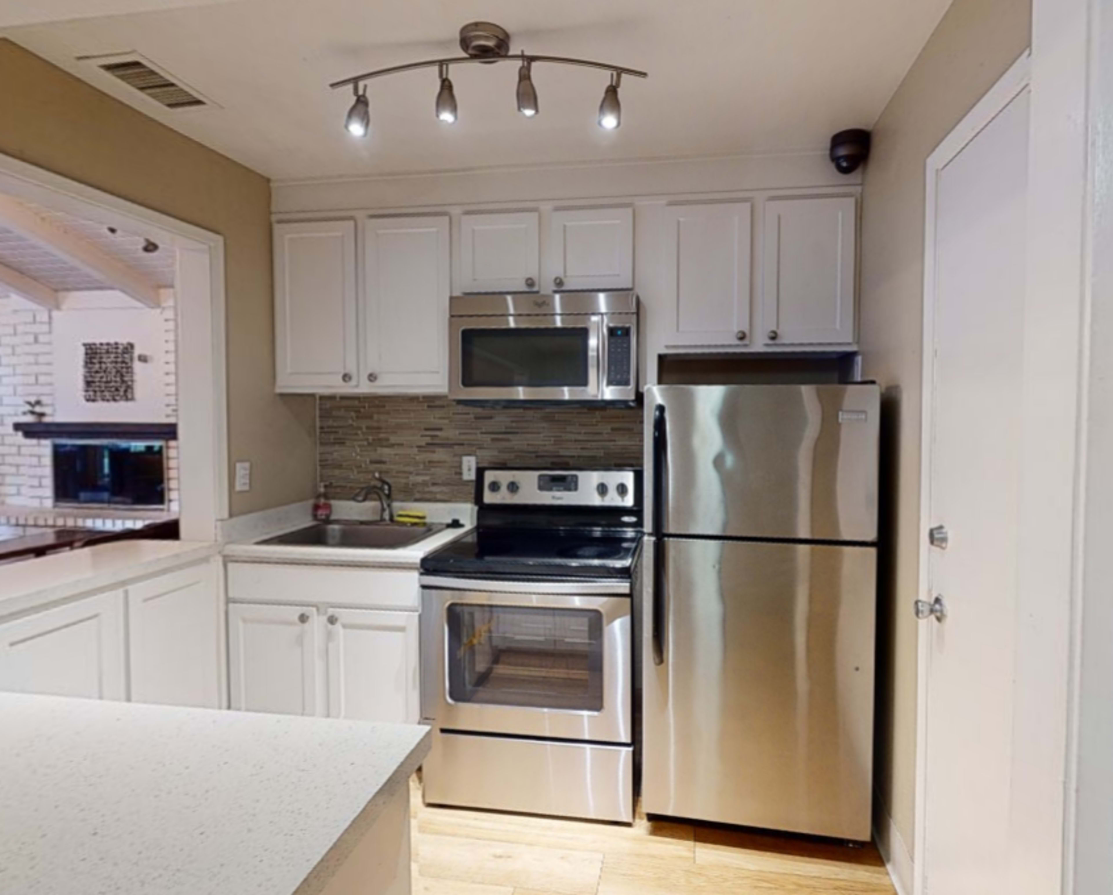 A compact kitchen features stainless steel appliances, including a microwave, oven, and refrigerator, with white cabinetry and a light-colored countertop.