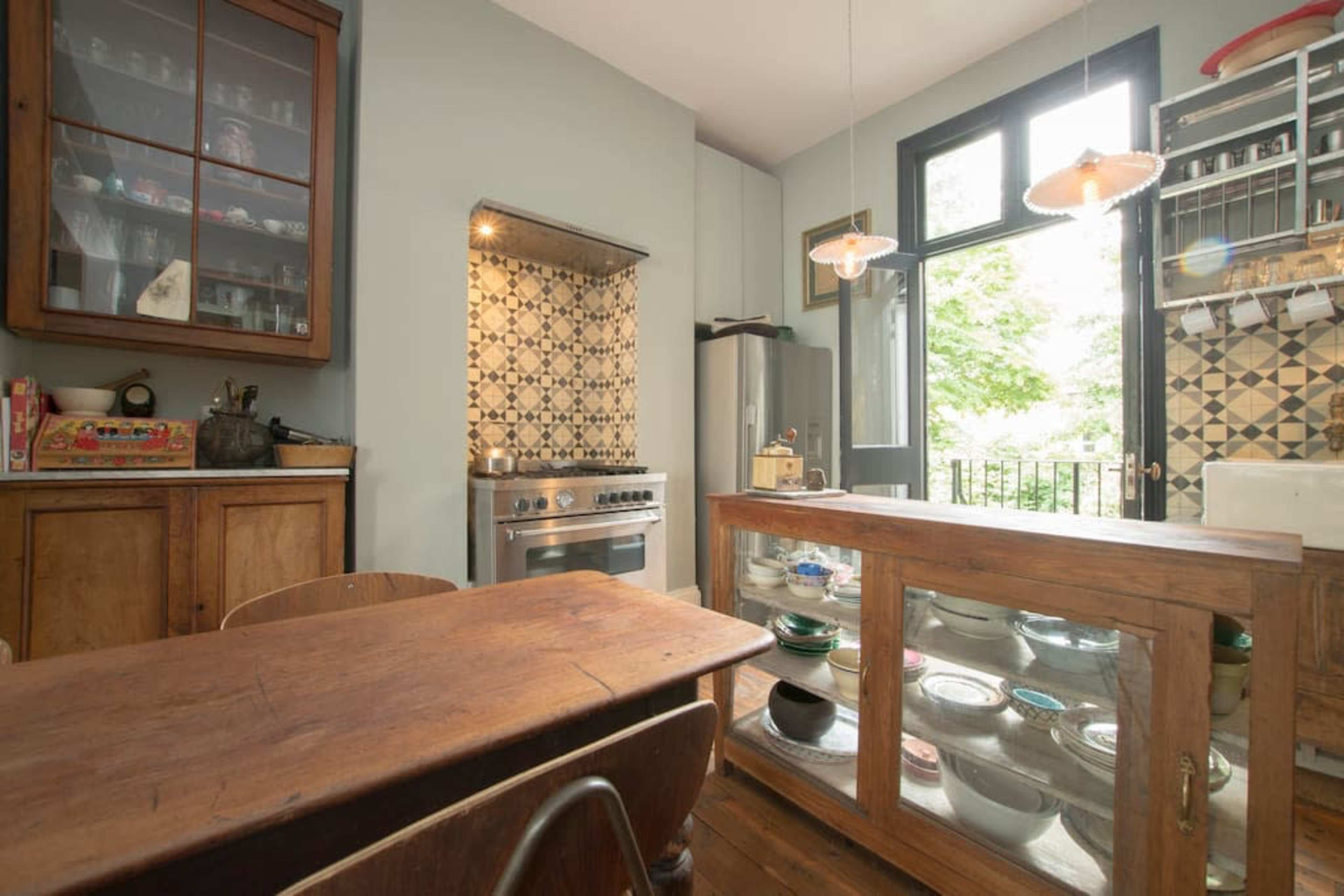 A kitchen features wooden furniture, a stainless steel stove, and large windows overlooking greenery.