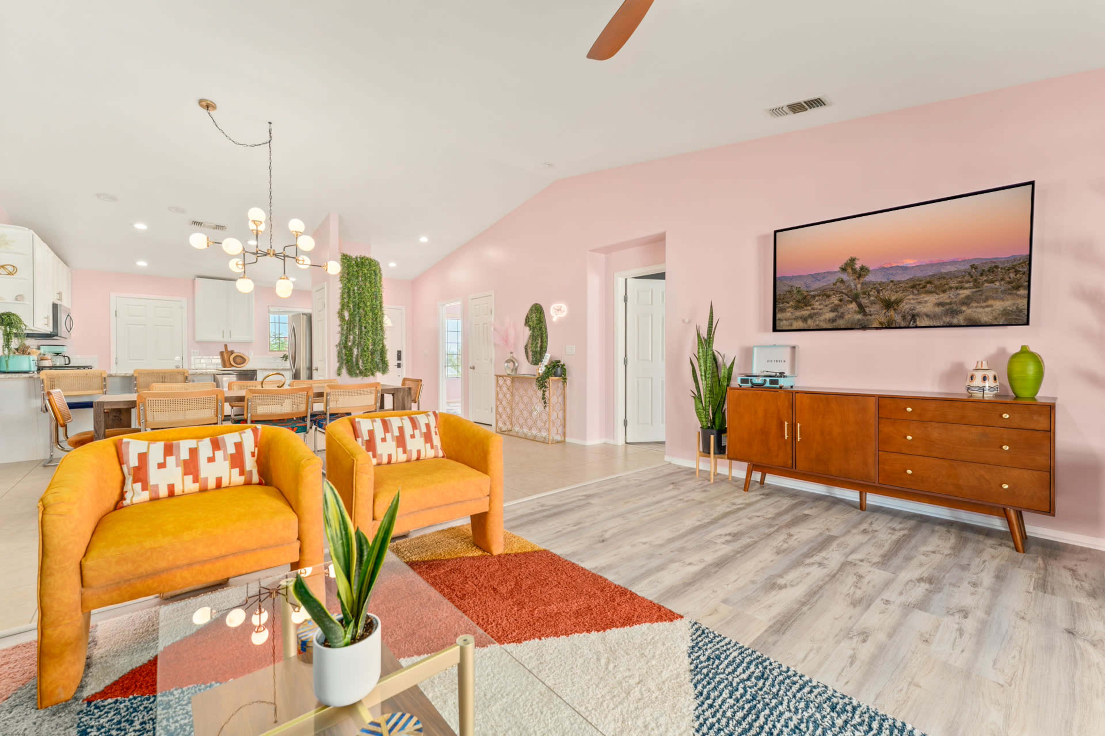 The image shows a bright living area featuring two orange chairs, a wooden sideboard, a large TV mounted on a pink wall, and a dining area in the background.