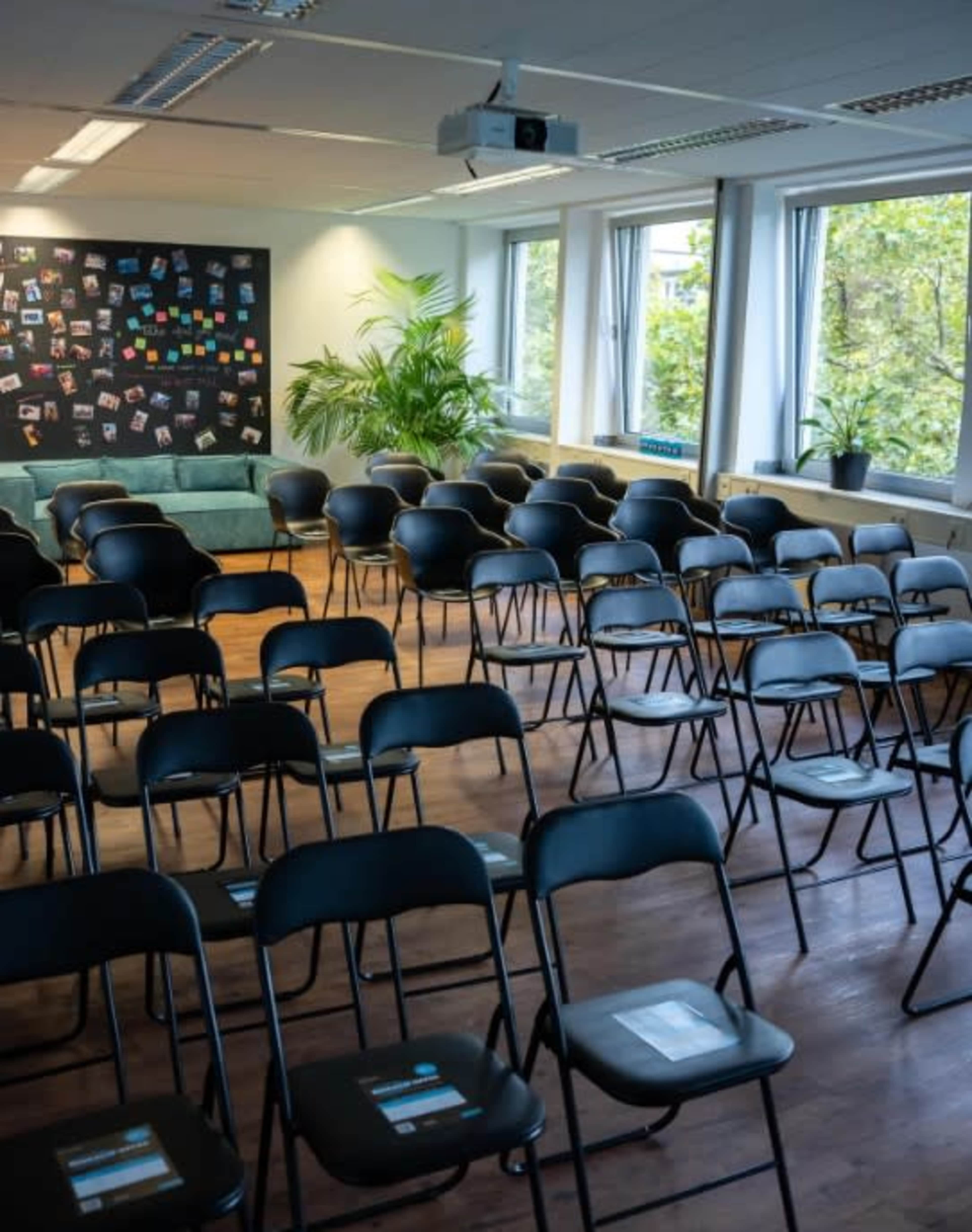 The image shows a room with a seating arrangement of black chairs facing a presentation area, complemented by a blackboard covered in photos and notes, along with a green couch and plants near the windows.