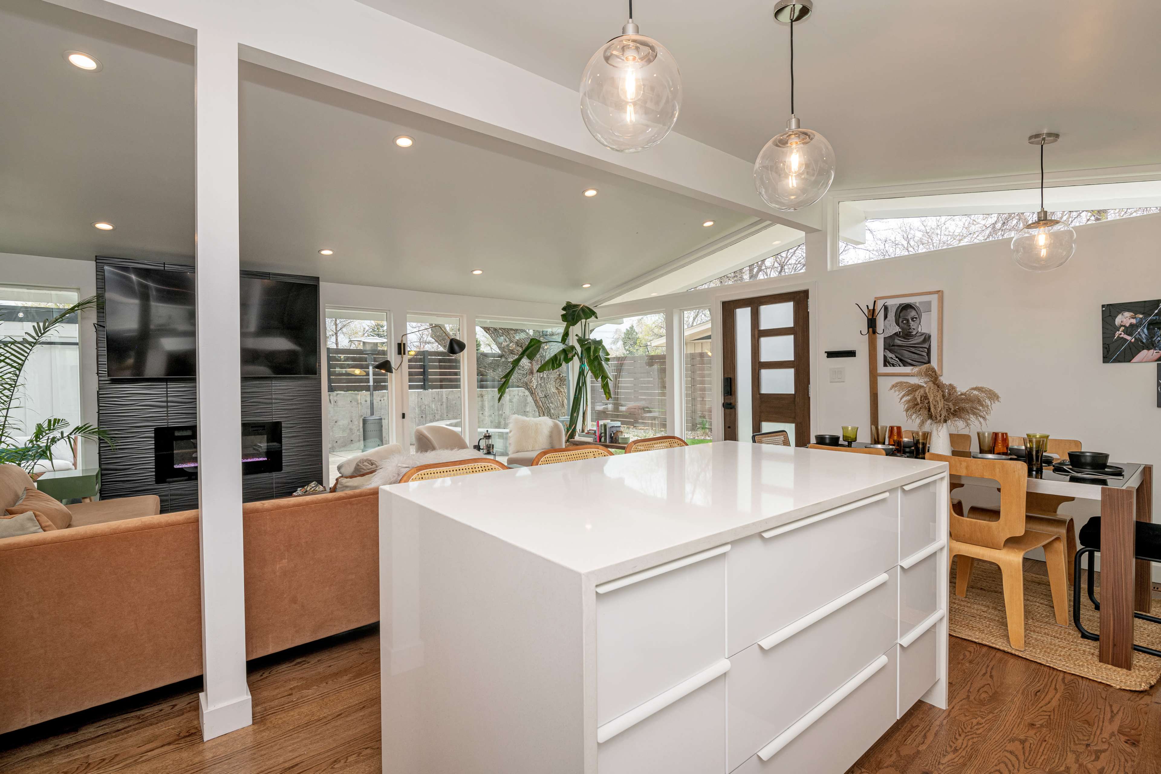 A modern kitchen with a white island, featuring pendant lighting and a seating area, connected to a living space with large windows.