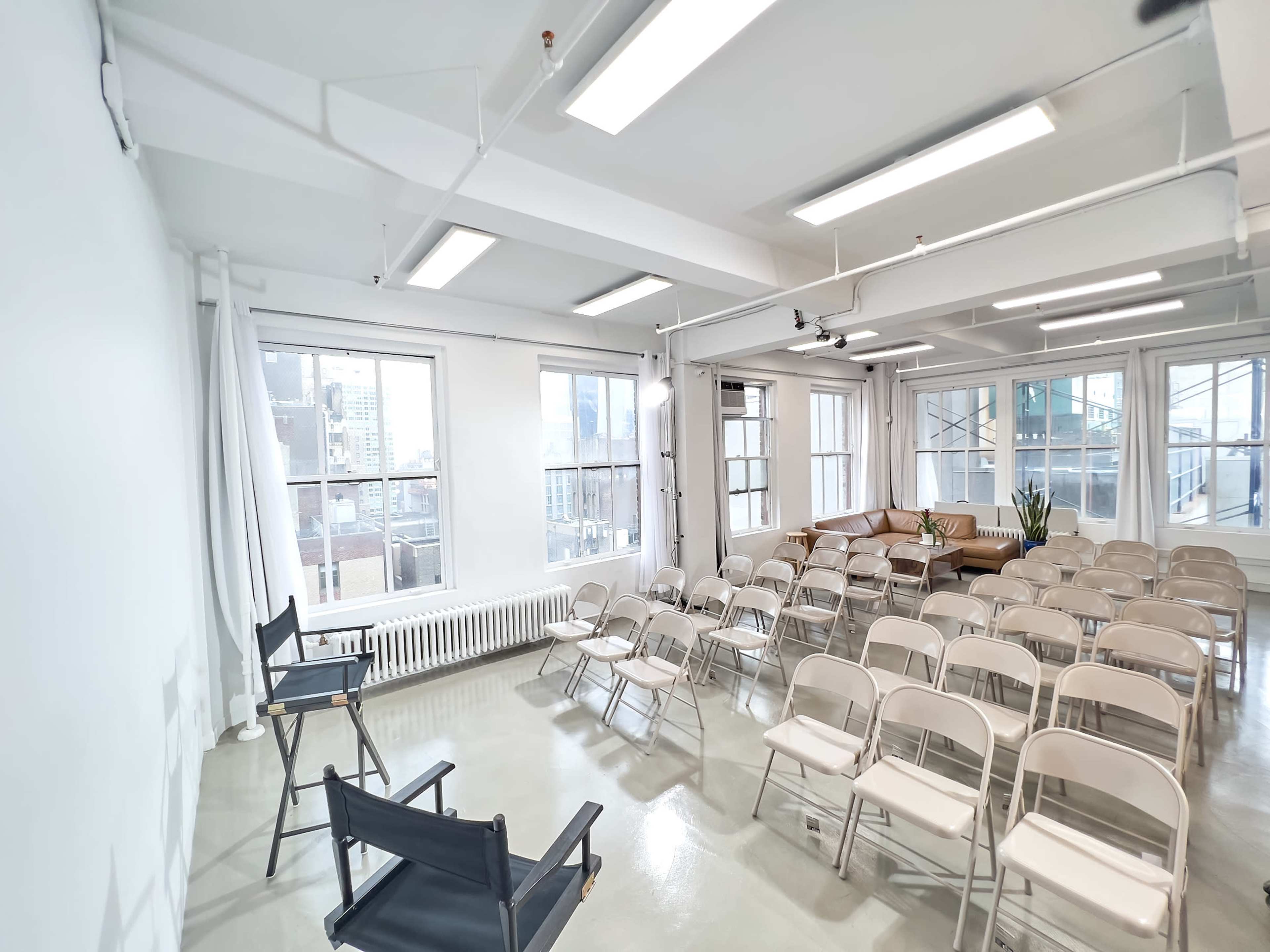 A brightly lit room with rows of beige chairs facing a blank wall and a small seating area with a couch and potted plant by the windows.