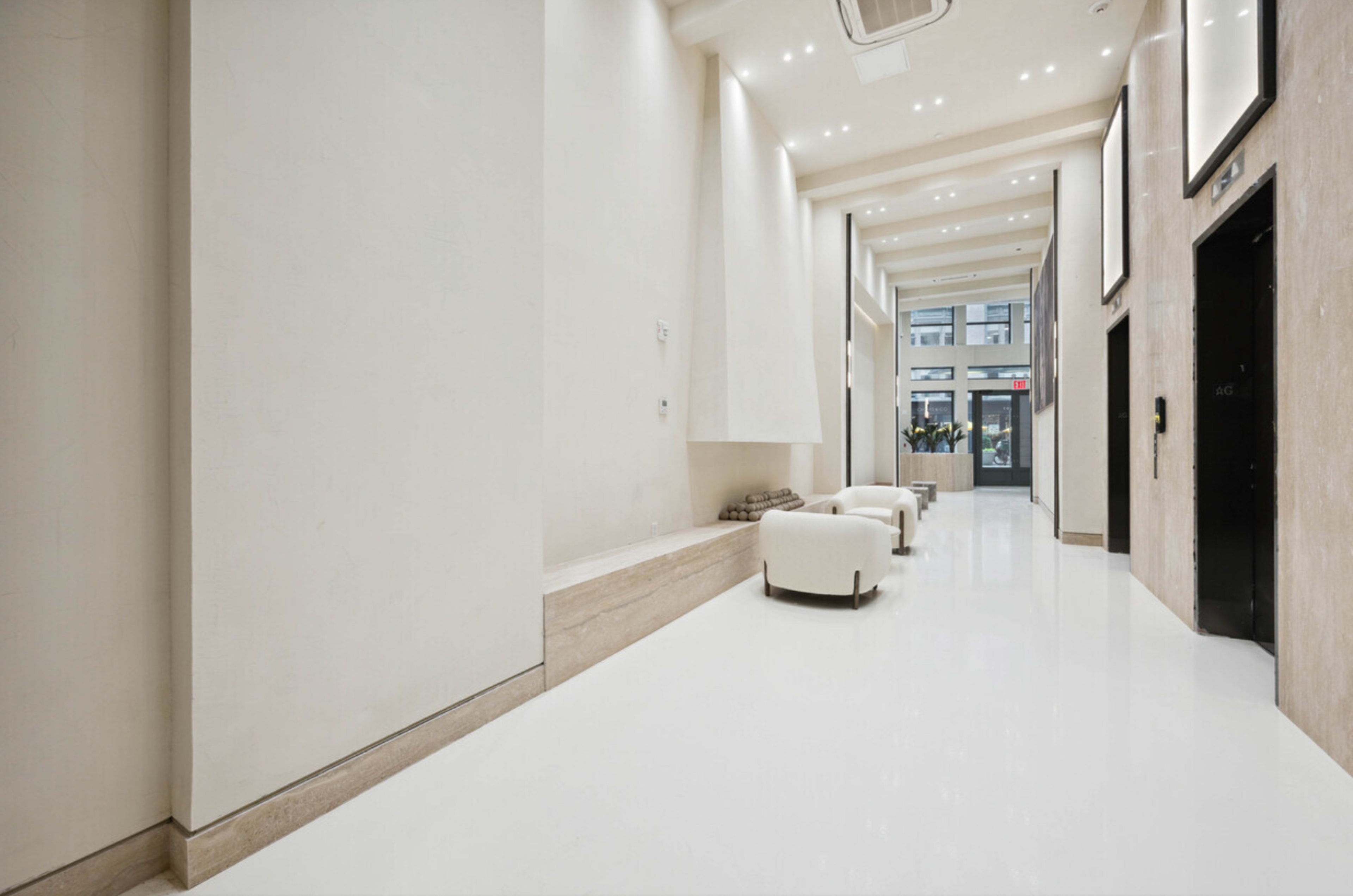 The image shows a spacious, well-lit hallway with white walls and a glossy floor, featuring two white chairs and a row of windows at one end.