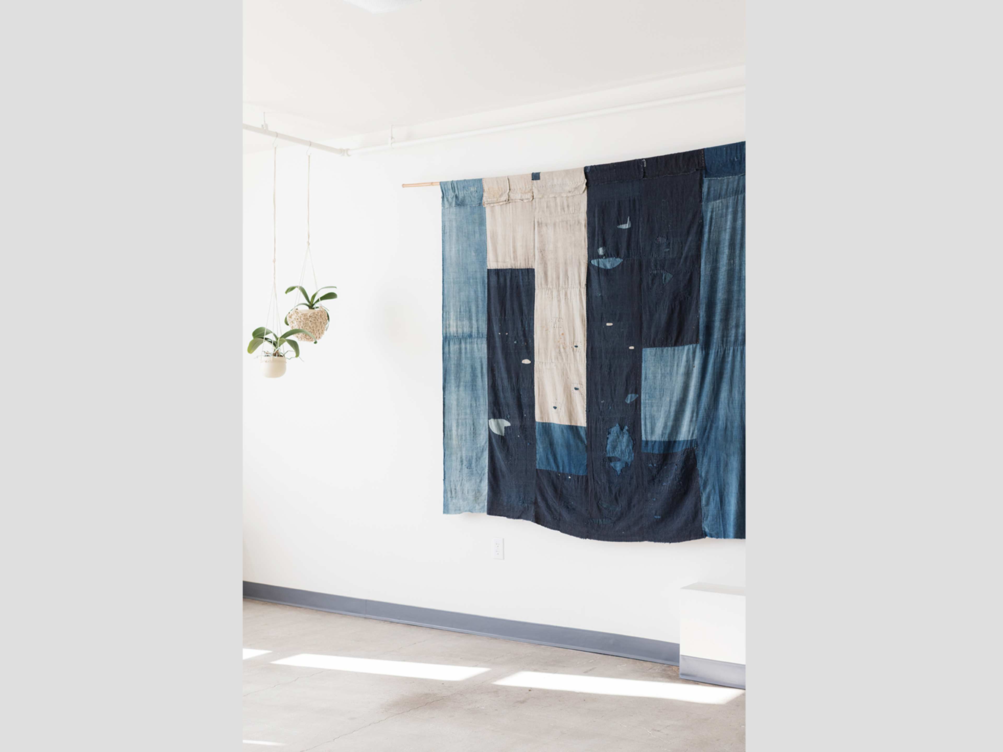 A wall hanging made of patchwork fabric in various shades of blue, displayed in a minimalist room with a plant in a hanging pot.