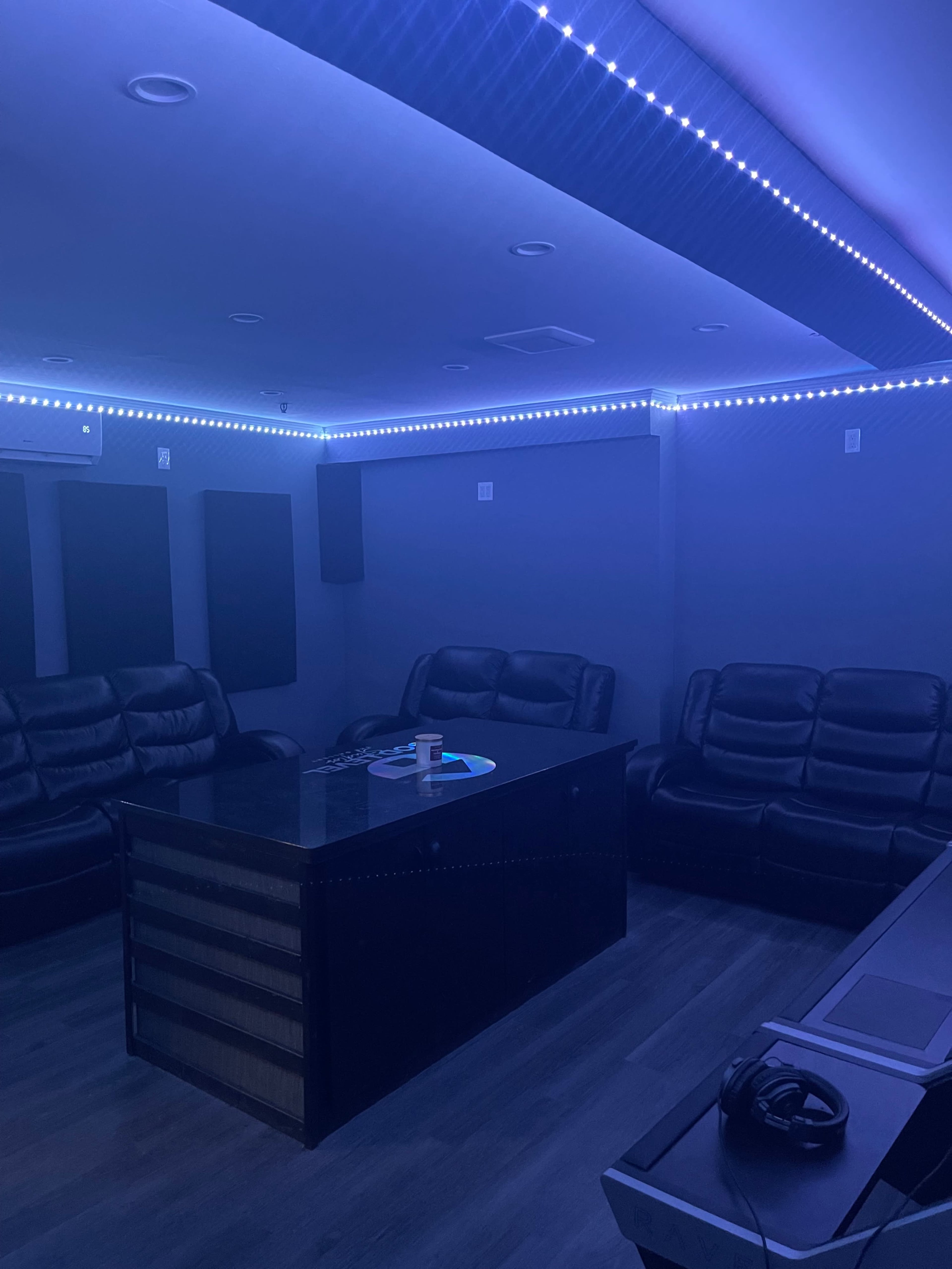 A dimly lit room with blue LED strip lighting, featuring black leather couches and a central dark wooden table.