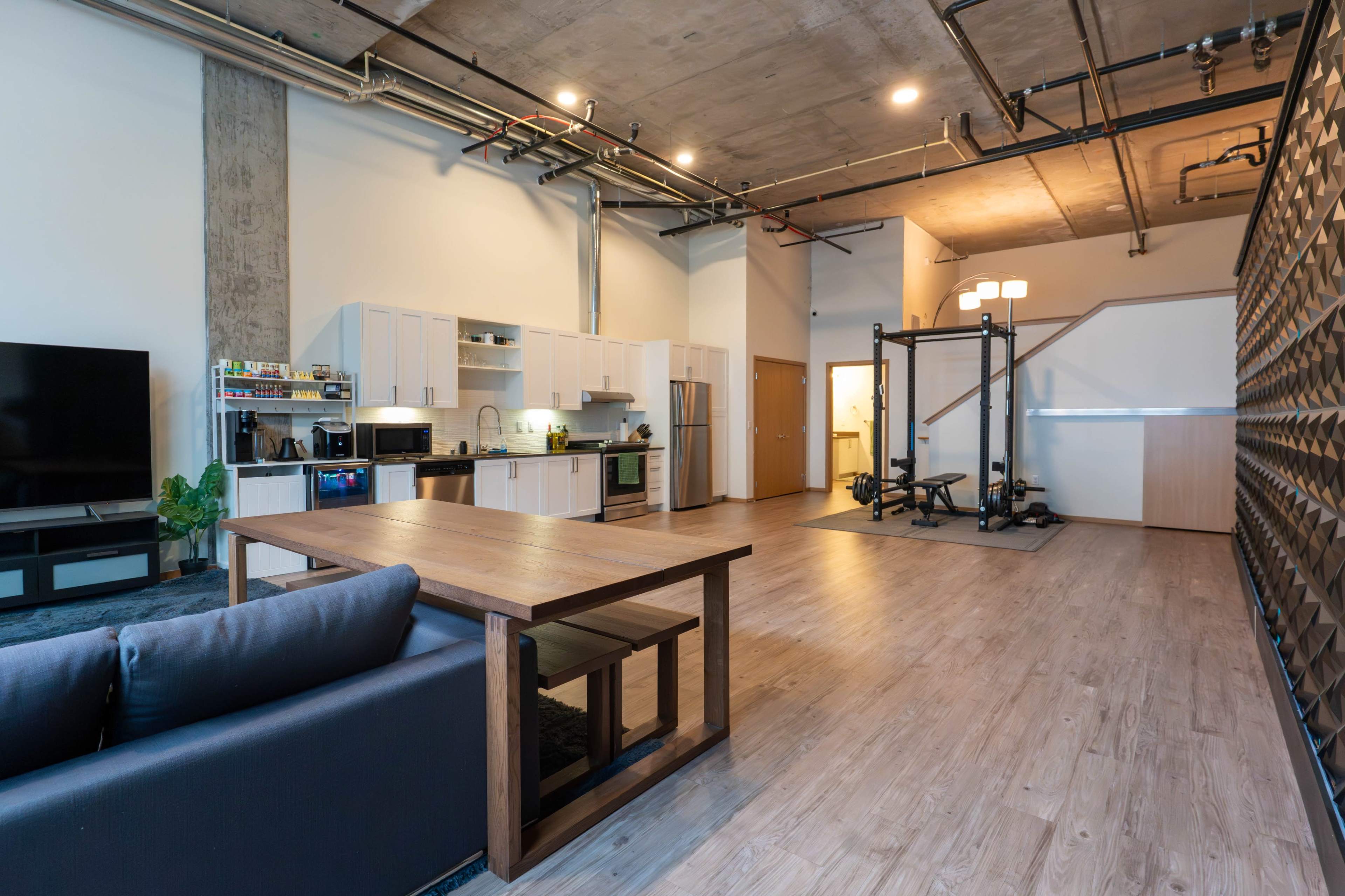 The image shows a spacious, modern studio apartment featuring a kitchen area, dining table, living space, and a small home gym setup.